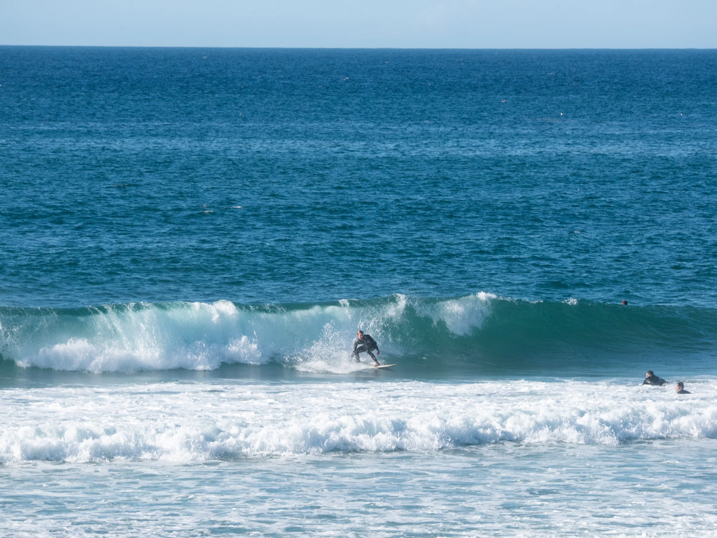 A person surfing on a wave at the beach, with others in the water nearby, in front of the ocean and clear sky.