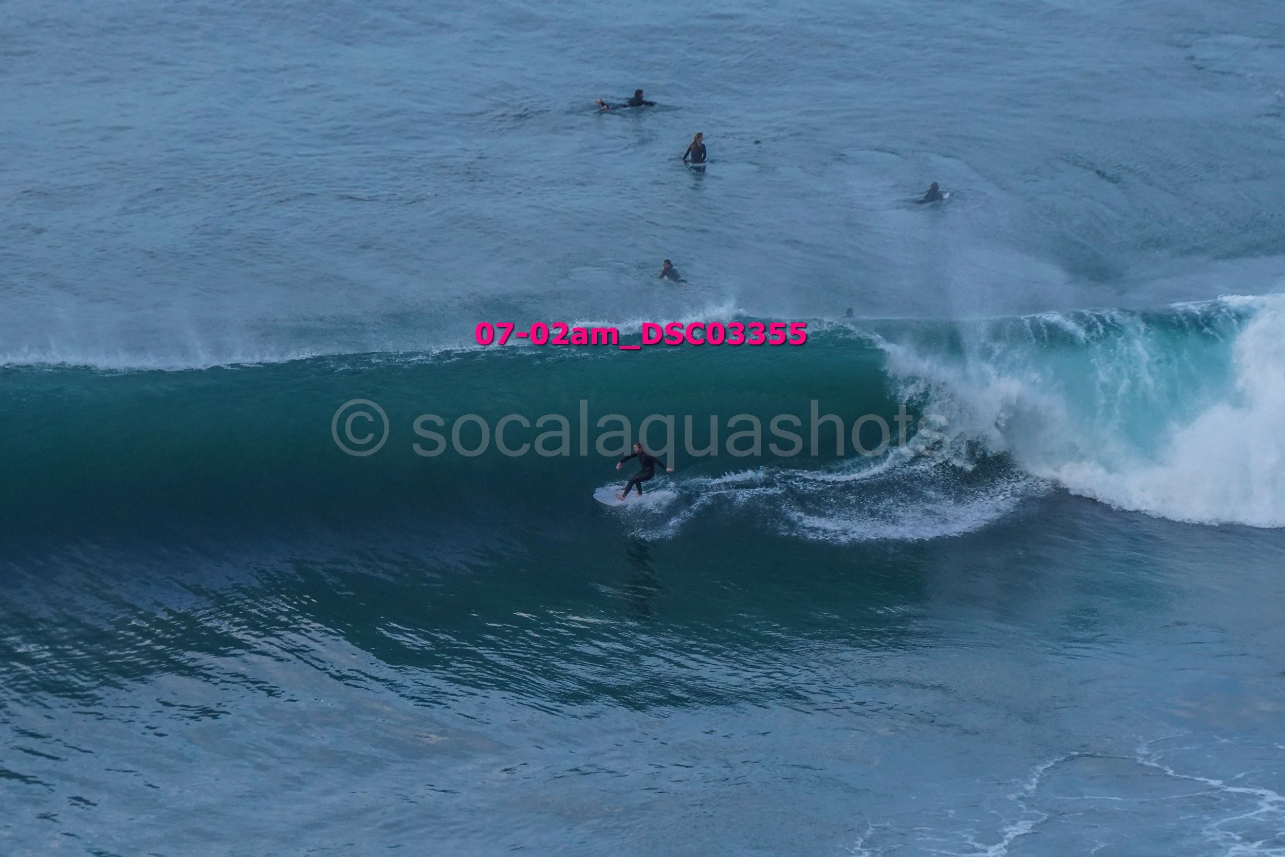 A surfer rides a wave with several people swimming in the background in the ocean.