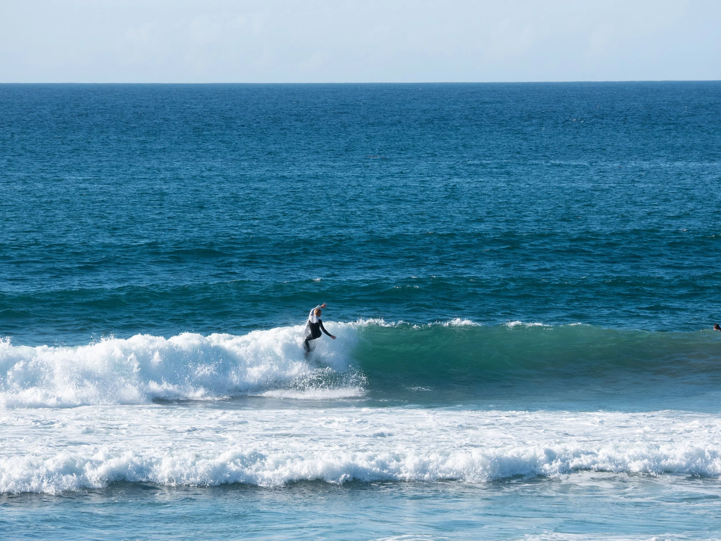 A person surfing on a small wave in the ocean with a clear blue sky above.