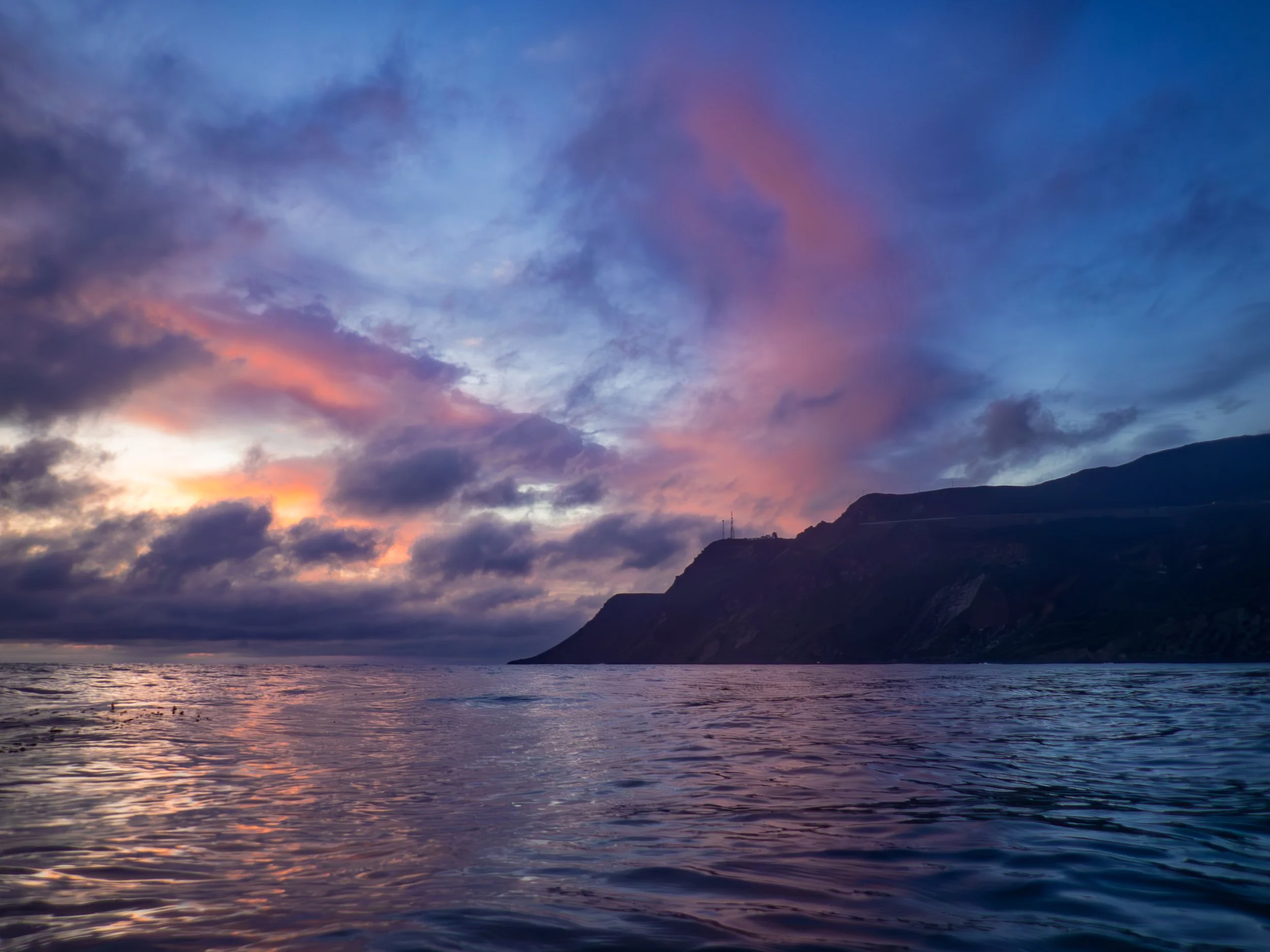 Sunset over the ocean with a mountain in the background, colorful pink, purple, and blue clouds in the sky.