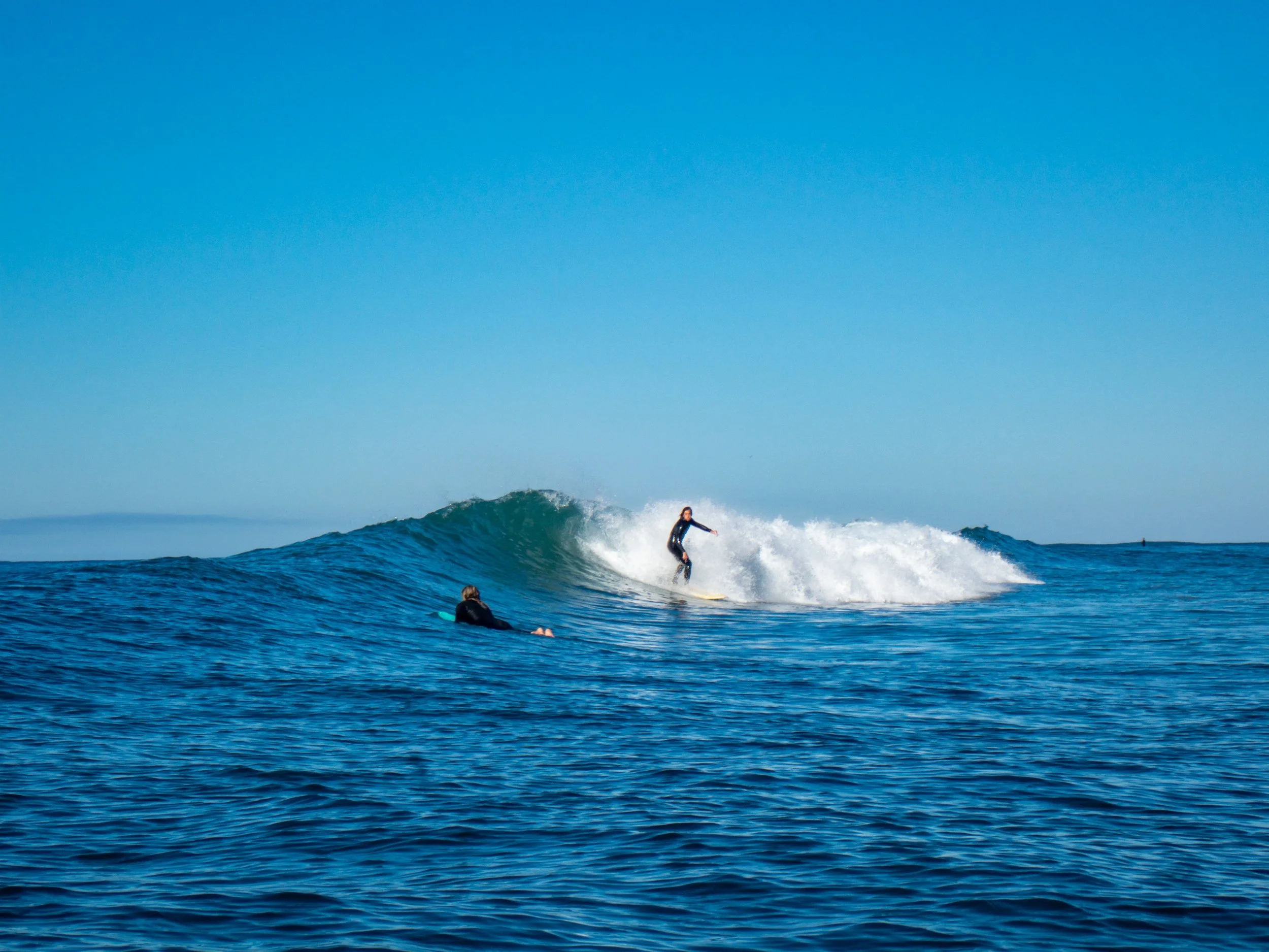 A person surfing on a wave in the ocean with another person floating nearby under a clear blue sky.