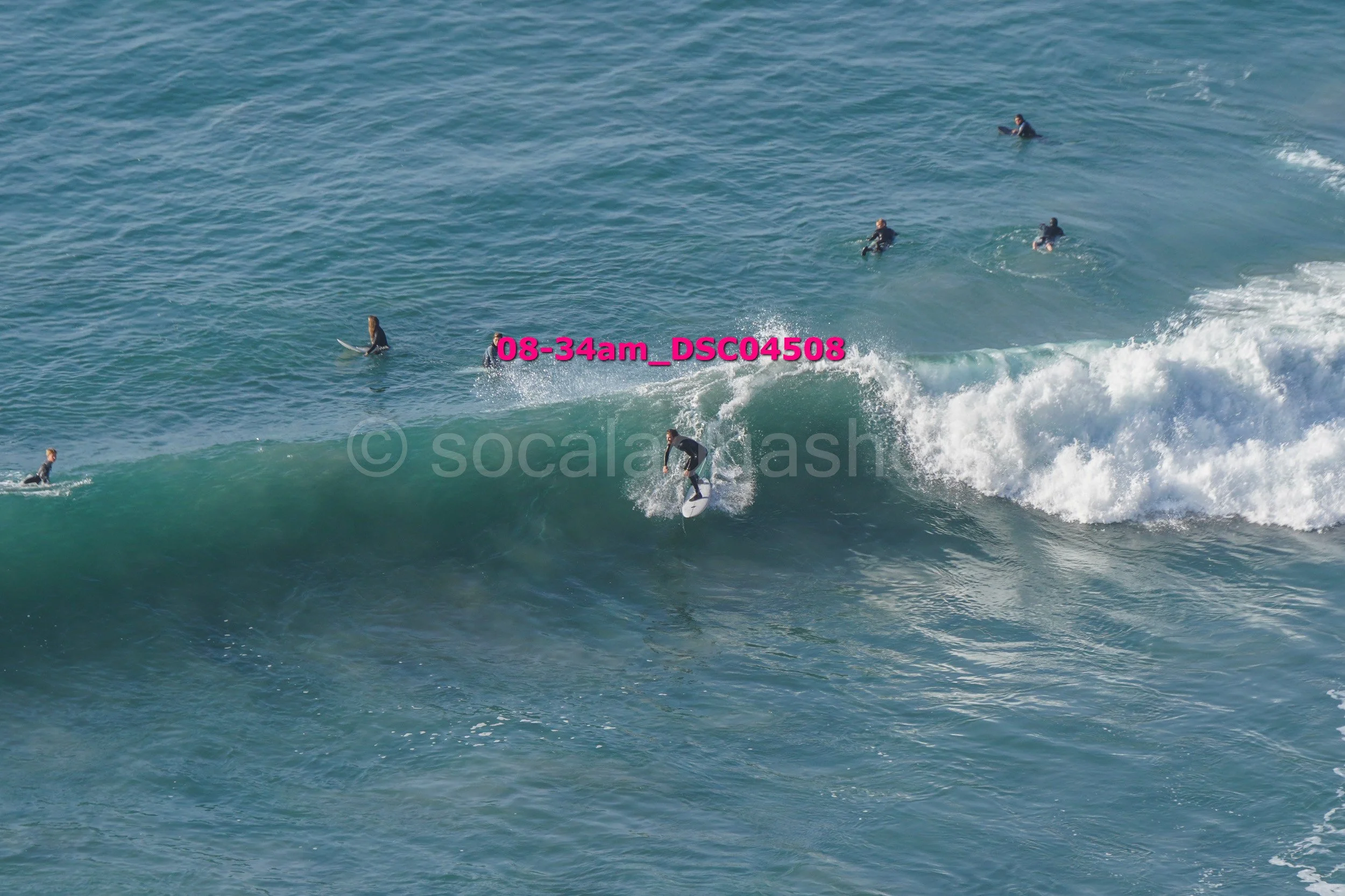 Surfer riding a wave with several people in the water around him, some holding surfboards.