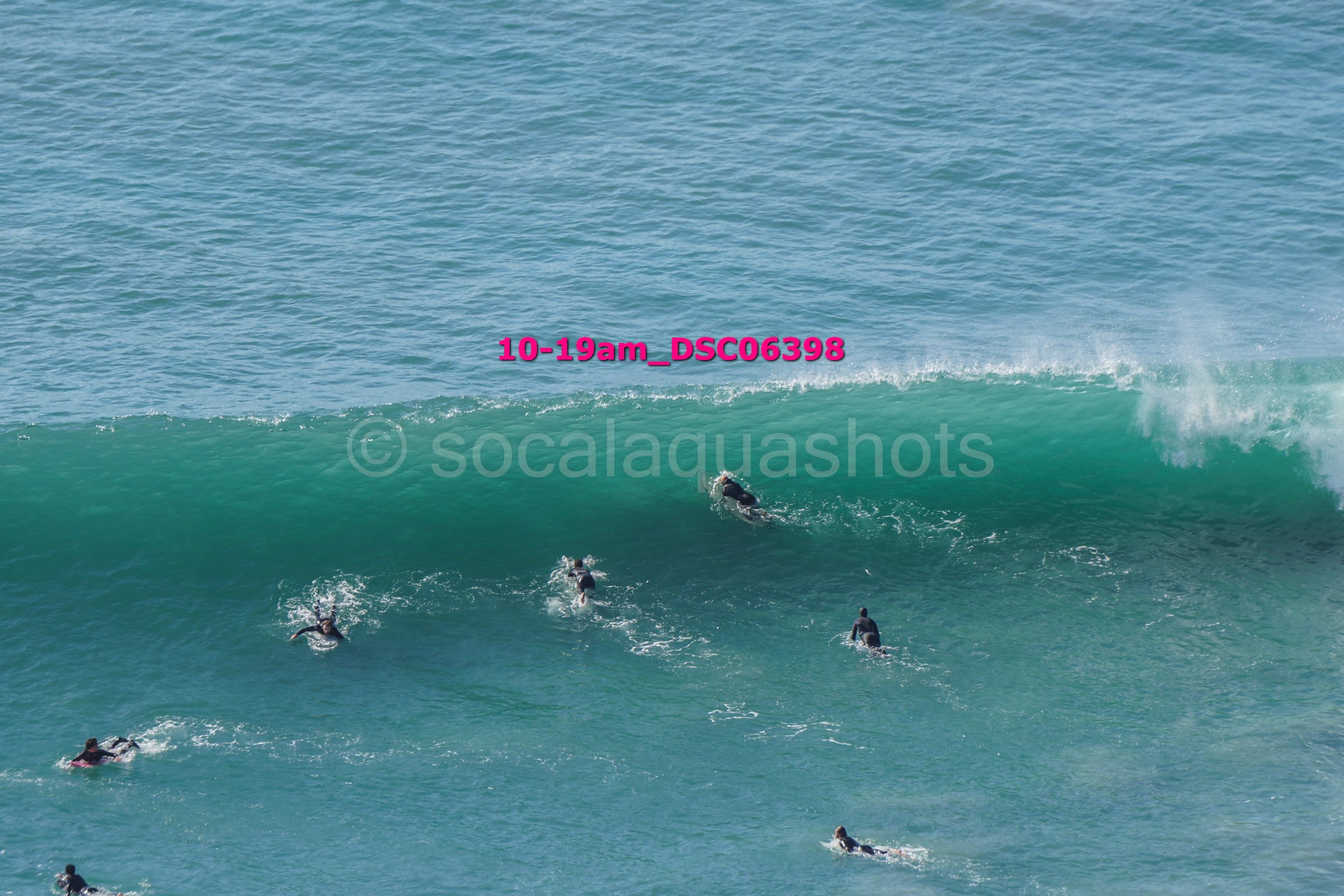Surfers riding a large blue-green ocean wave, with some surfers paddling in the water.