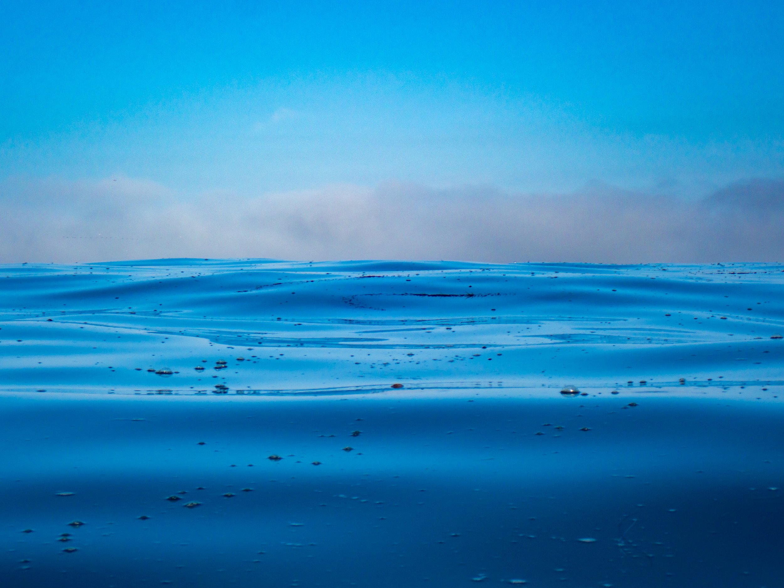 Wide view of calm blue water with small ripples, under a blue sky with some clouds on the horizon.