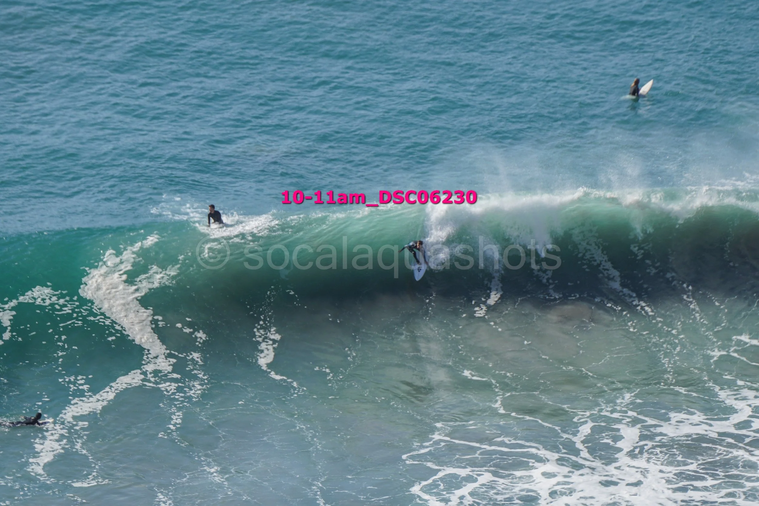 Surfer riding a large wave with two other surfers in the water and a paddleboarder in the background.