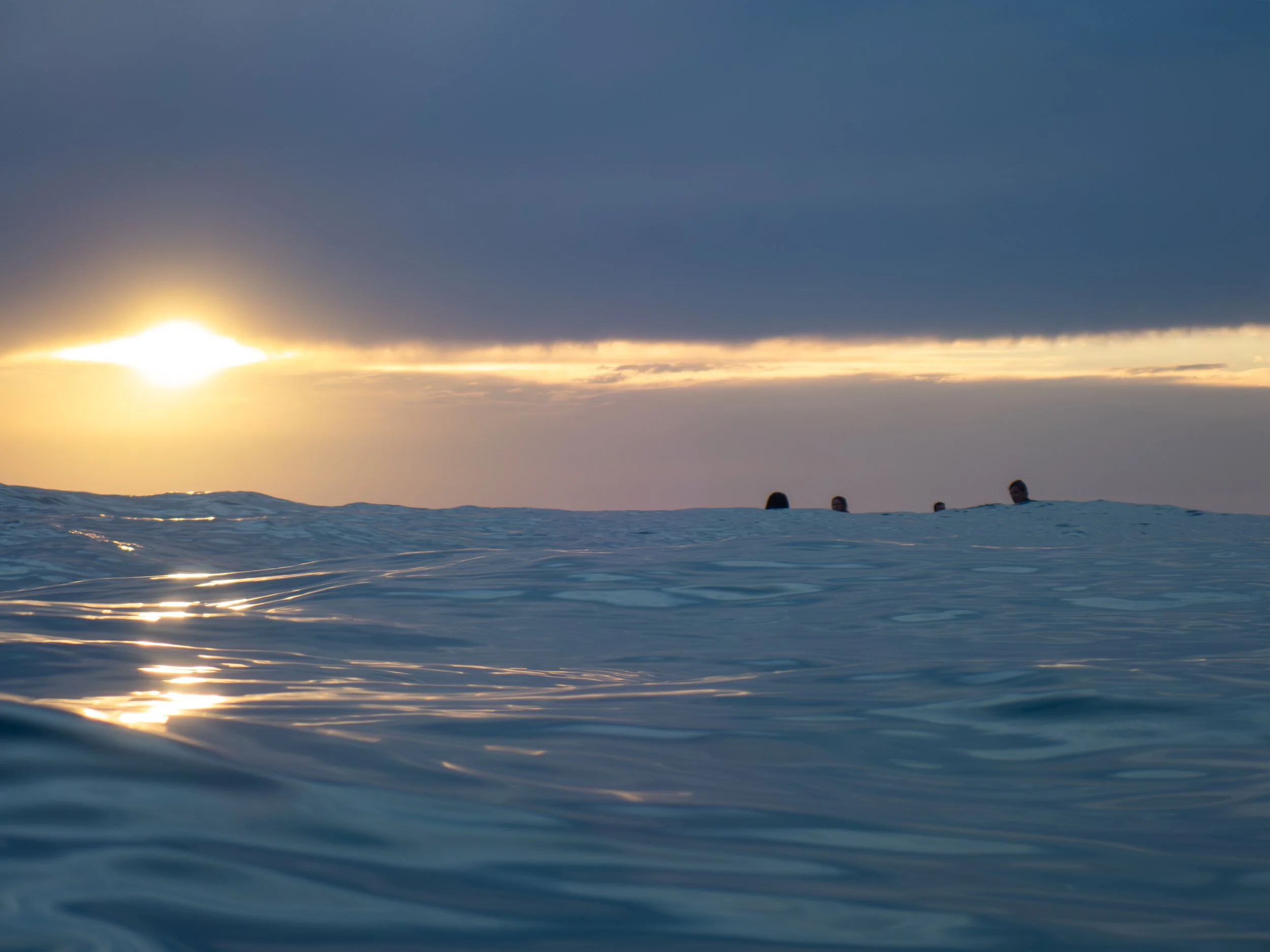 Overhead view of the ocean at sunset, with four people partially visible in the distance on the water's surface, under a partly cloudy sky.