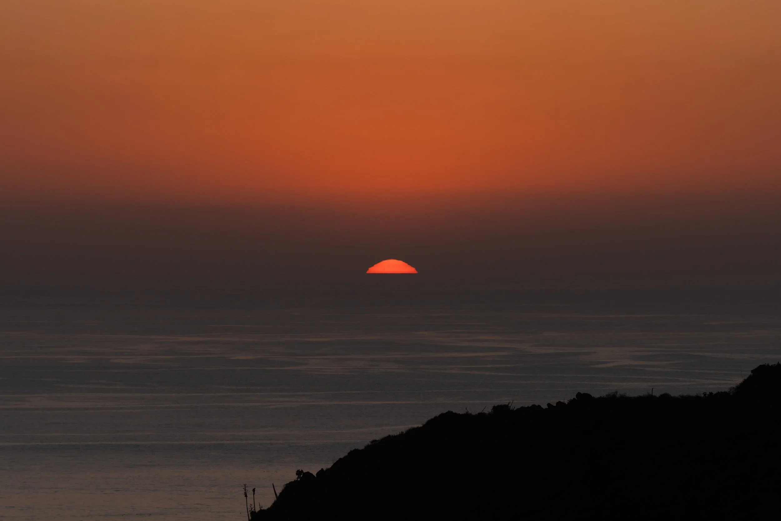 Sunset over the ocean with a dark silhouette of a hill in the foreground, and a partially visible sun near the horizon.