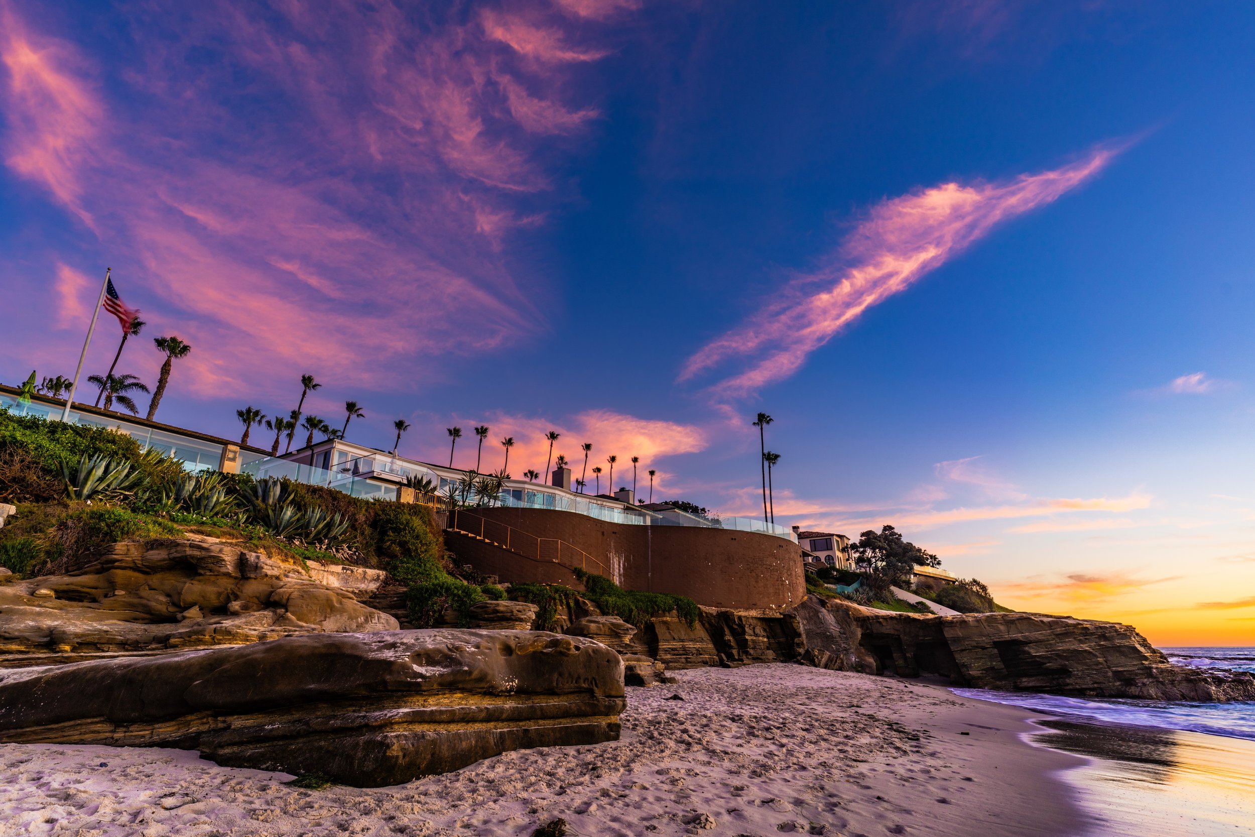A beach at sunset with rocky shoreline, sandy beach, and a house on a hill with tall palm trees and an American flag.