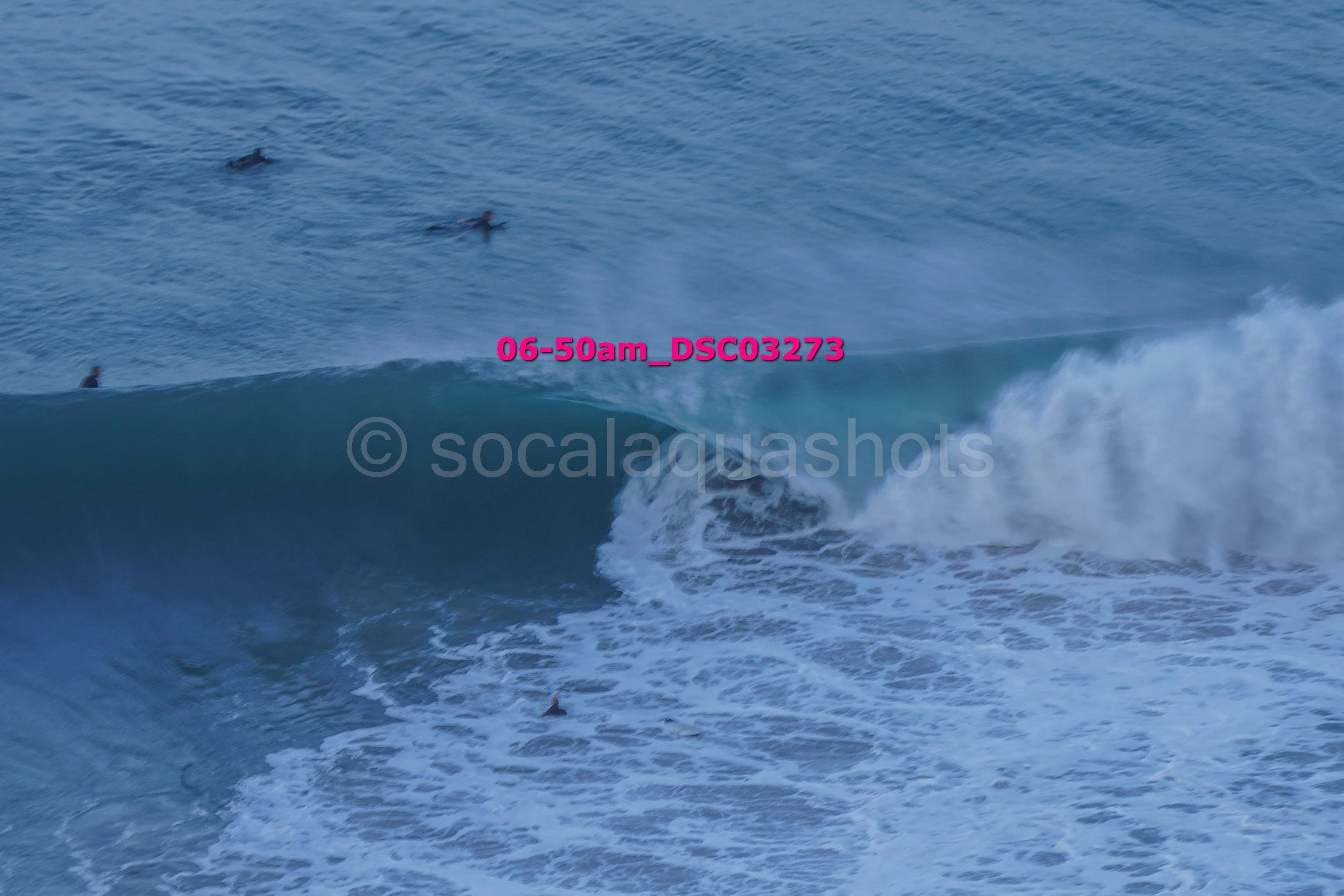 Several surfers in the ocean, with one riding a wave. The water is blue with white foam, and the scene appears early in the morning.