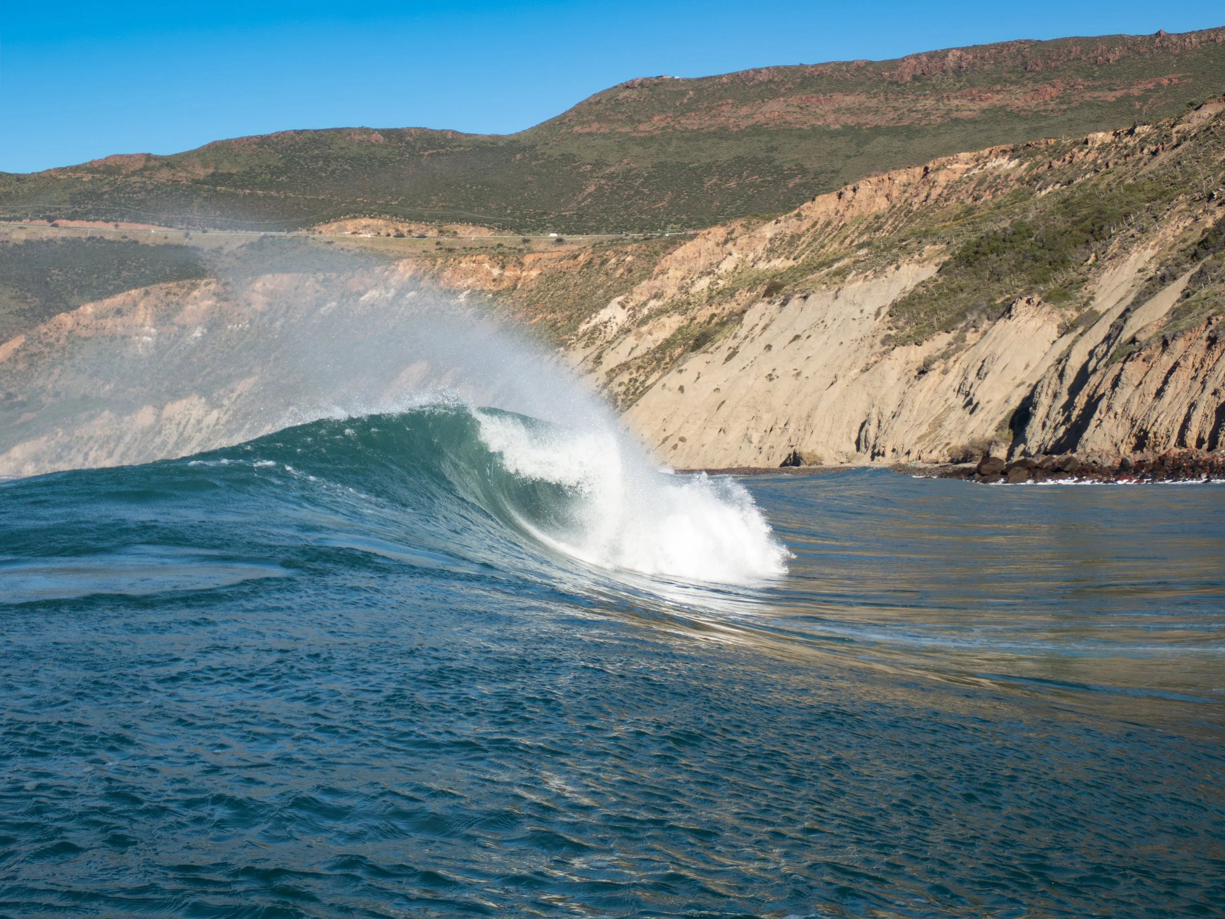 Ocean wave breaking near rocky coast with hills in the background on a sunny day.