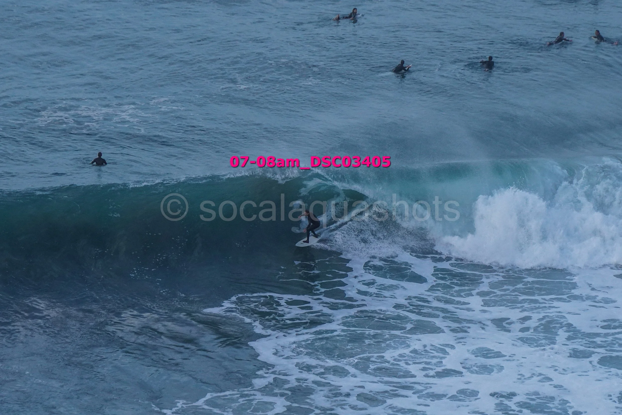 Surfer riding a large wave while several people swim in the ocean nearby.