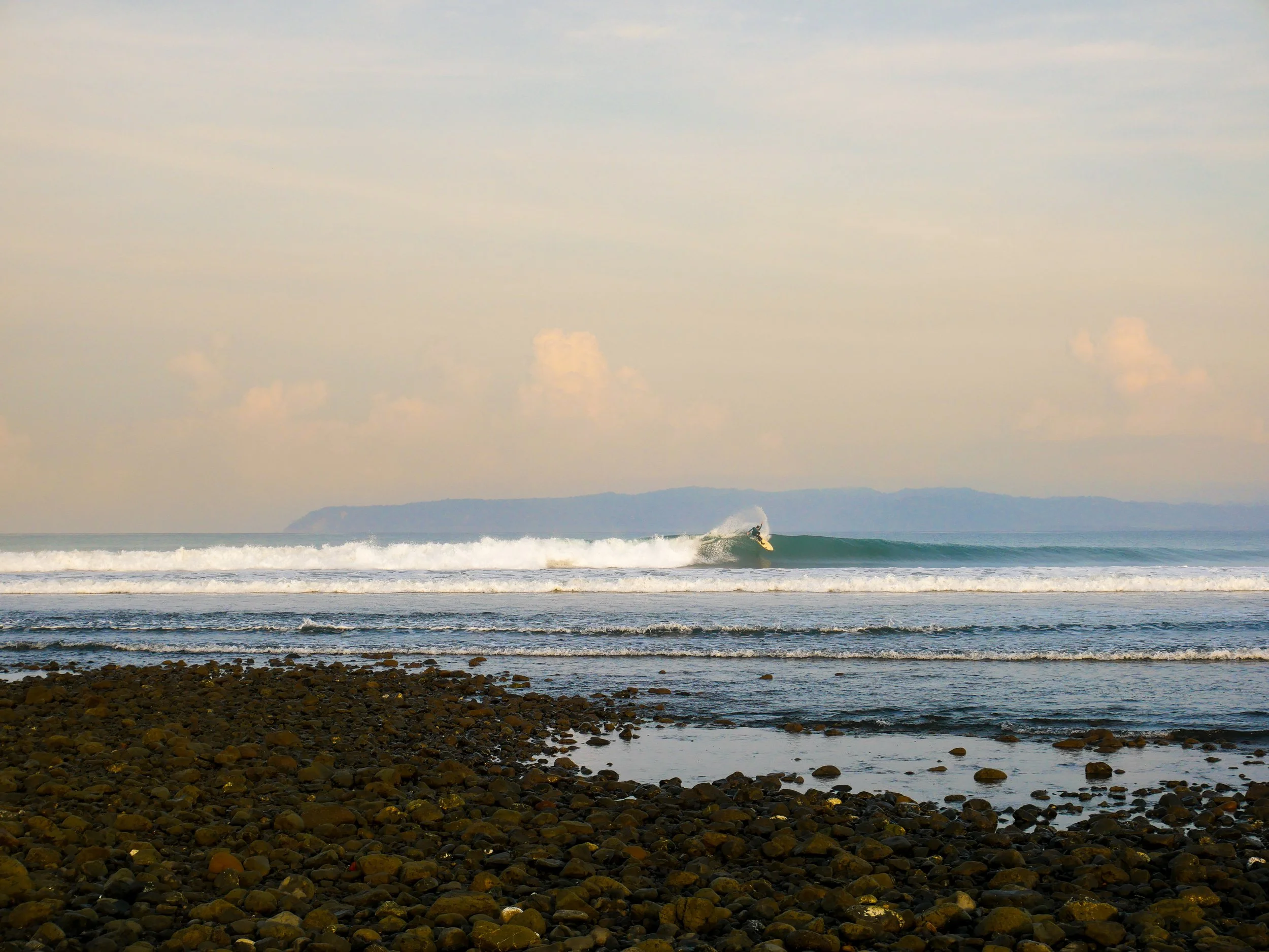 Surfer riding a wave at a rocky beach with distant hills, blue sea, and cloudy sky.
