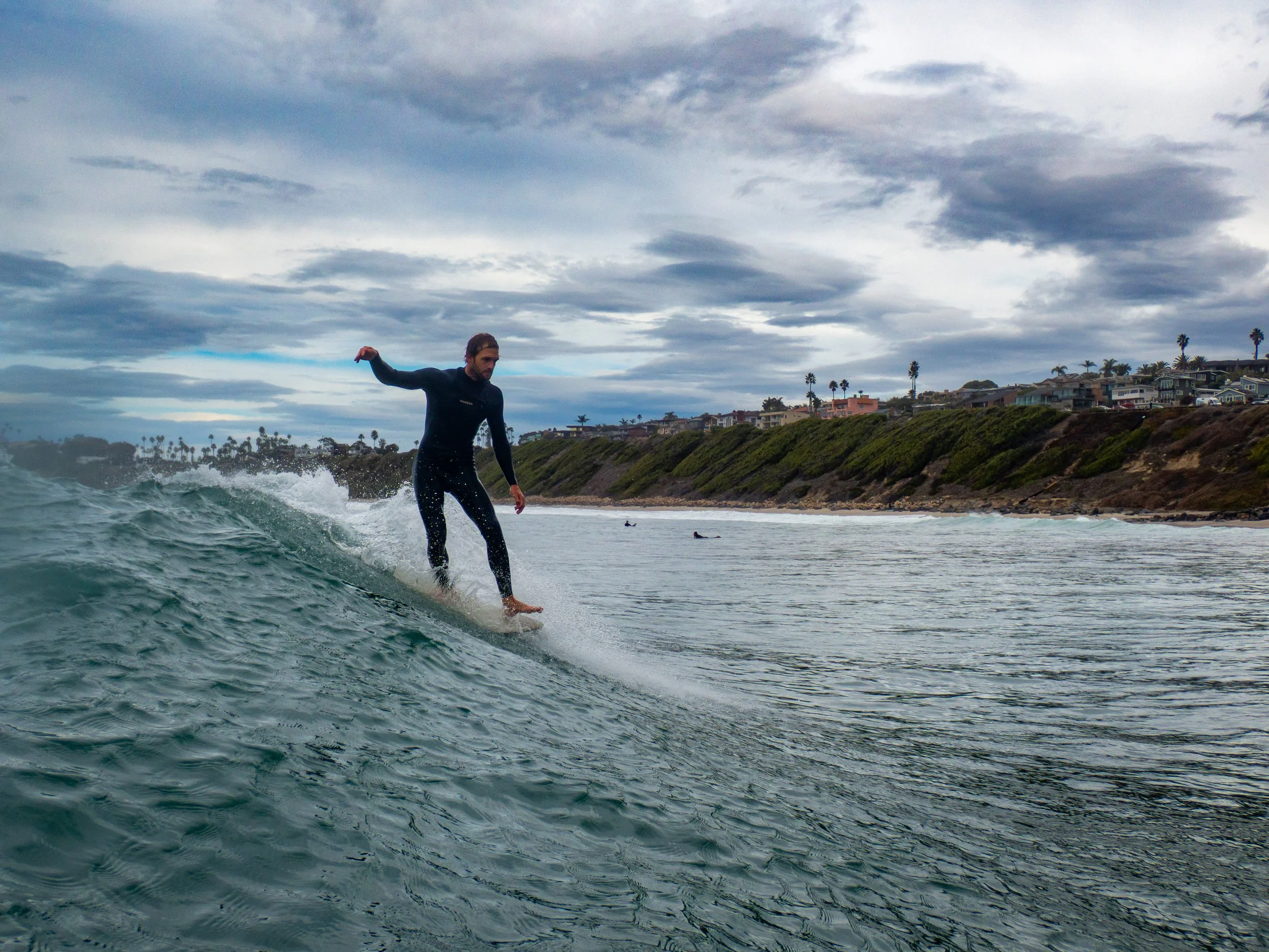 A man surfing on a wave near the shore with houses and palm trees in the background on a cloudy day.