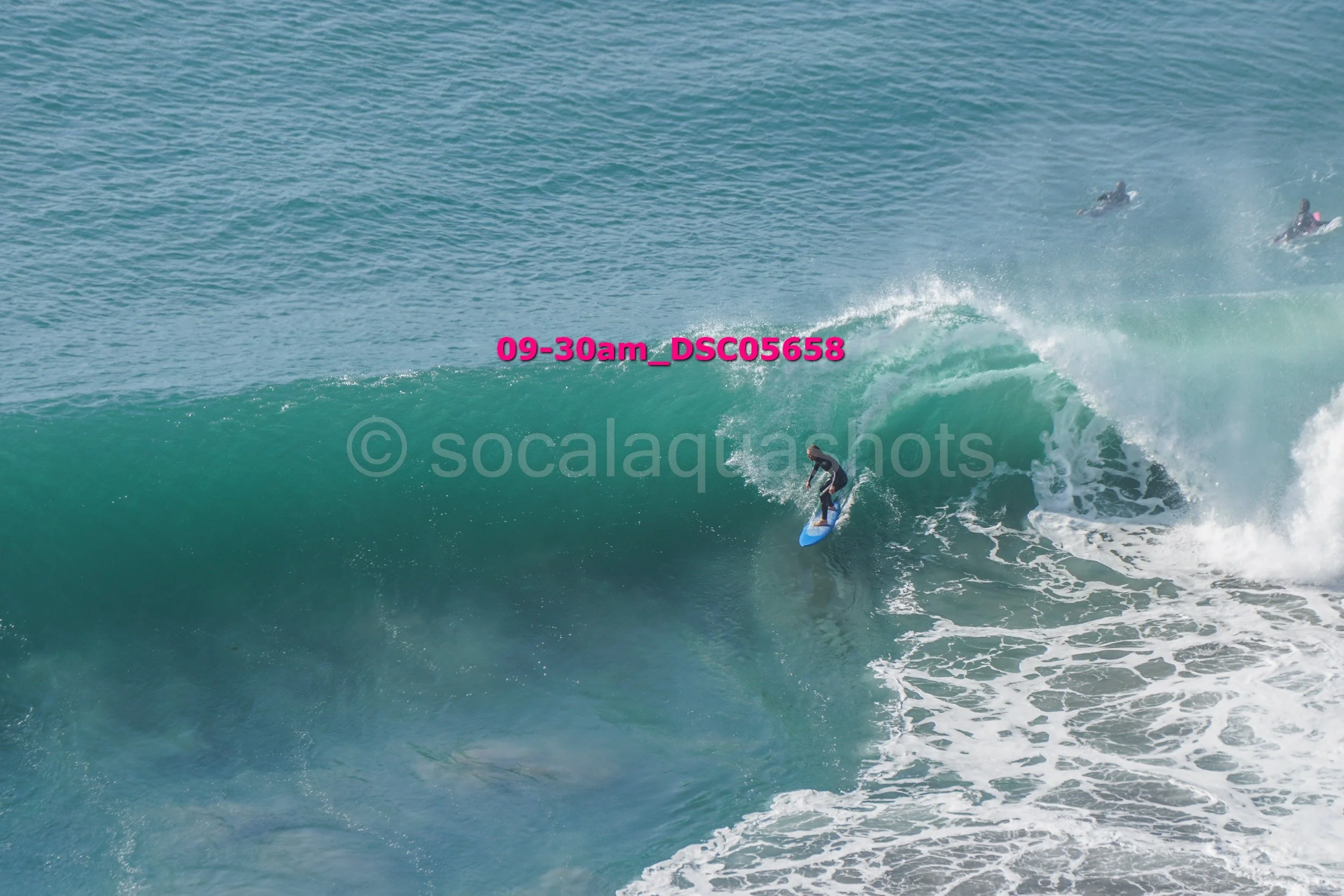 A person surfing on a large ocean wave with two other surfers in the background.