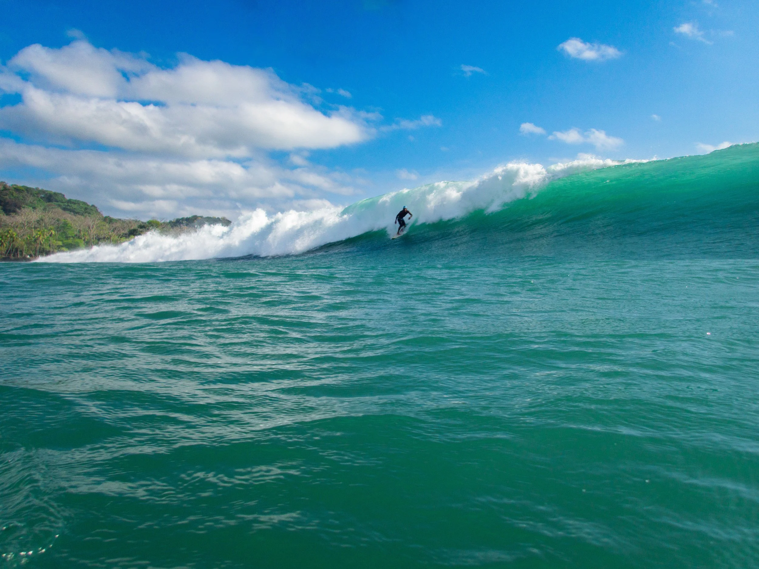 Surfer riding a large wave on a sunny day with a clear blue sky and sea in the foreground.