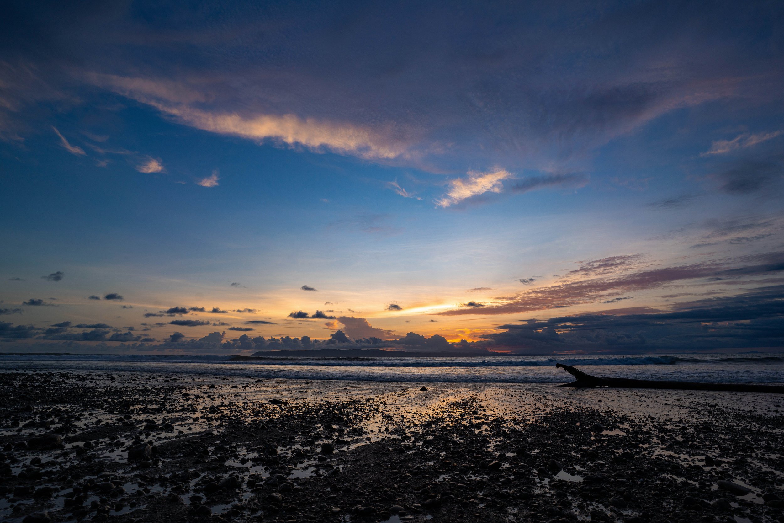 Sunset over a beach with rocks and driftwood, partly cloudy sky with pink and orange hues
