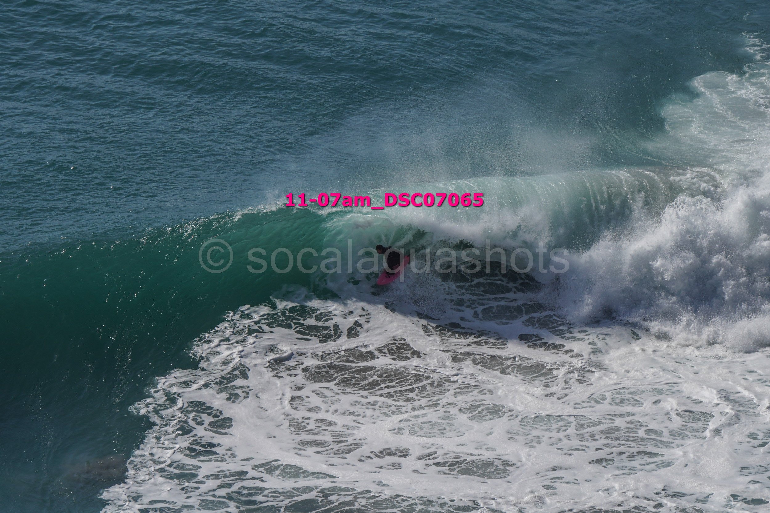 A surfer riding inside the barrel of a large ocean wave with white foam, visible despite the wave's greenish color.