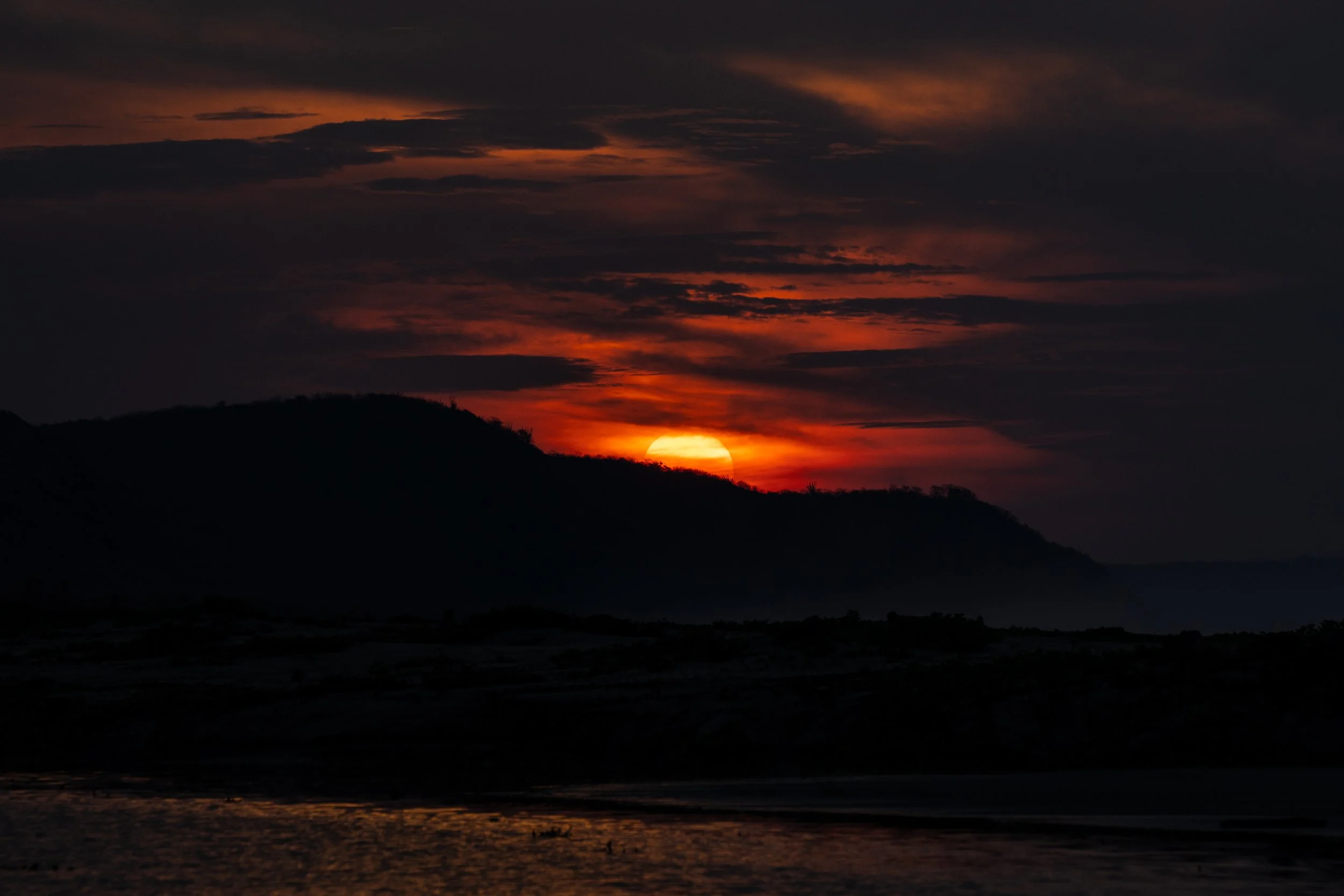 Sun setting behind mountains with a sky full of dark clouds and orange hues, reflected on water in the foreground.