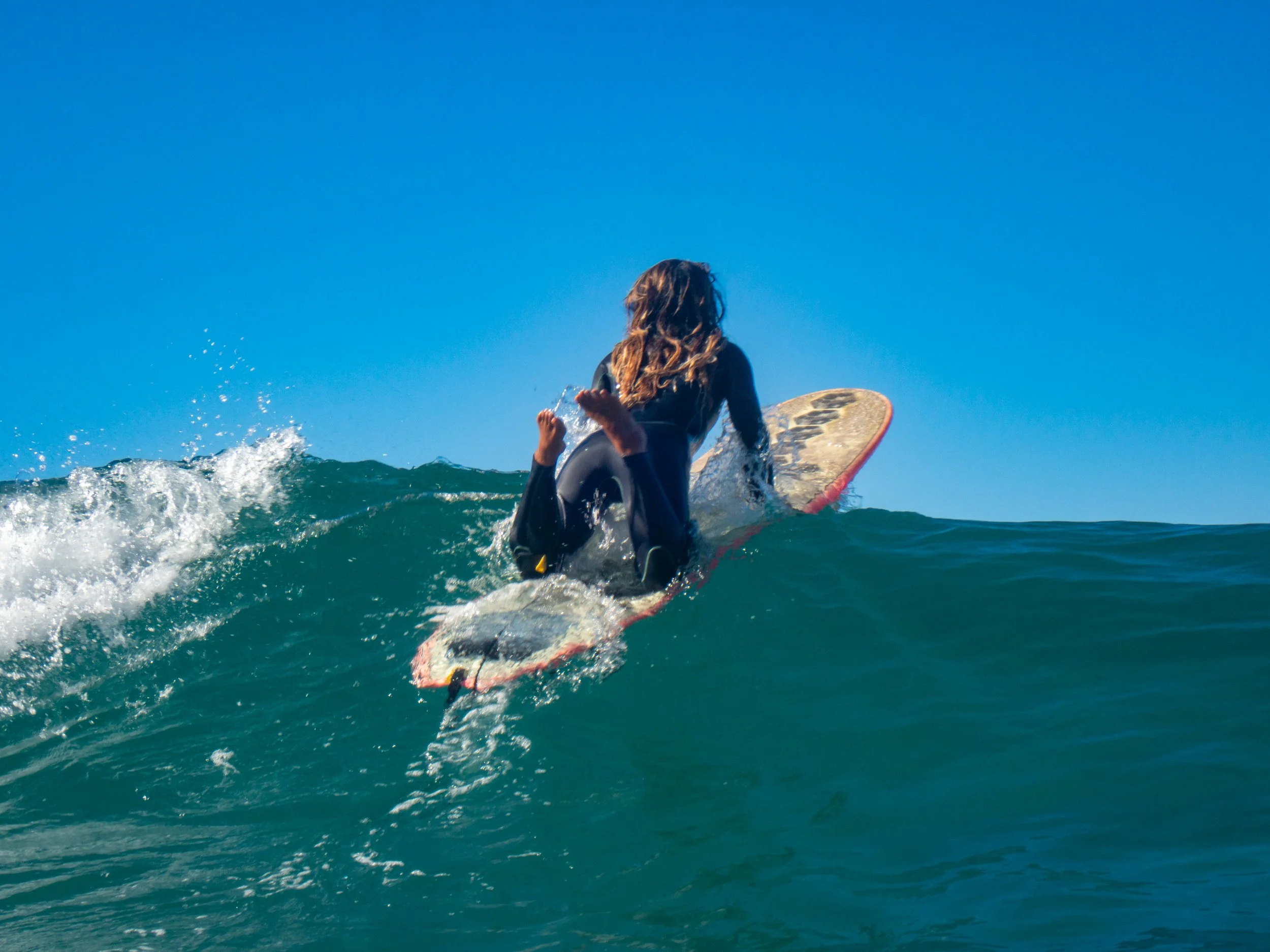 A person with long hair surfing on a wave with a clear blue sky in the background.