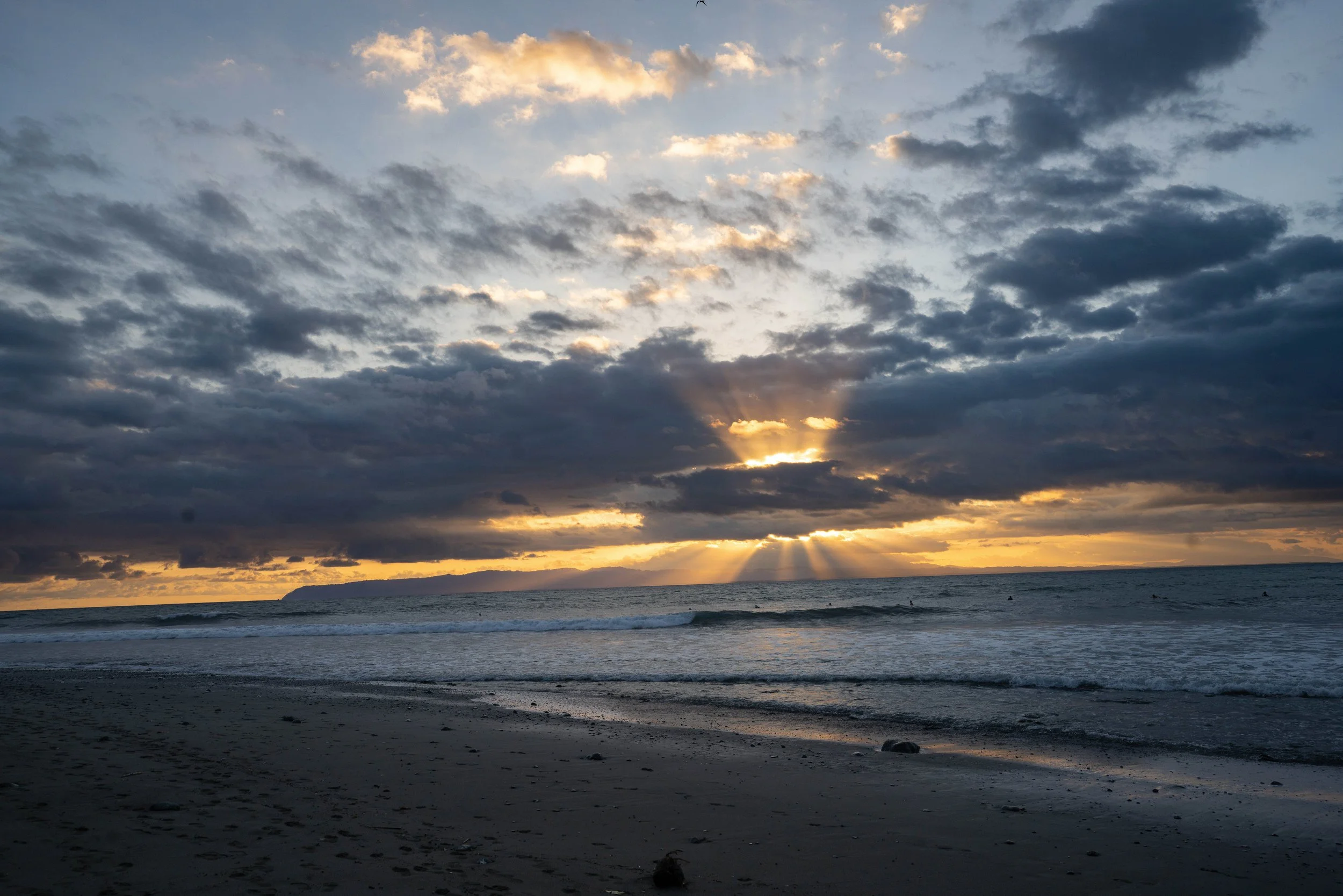 Sunset over a cloudy sky with the sun's rays breaking through clouds over a calm ocean and a sandy beach in the foreground.