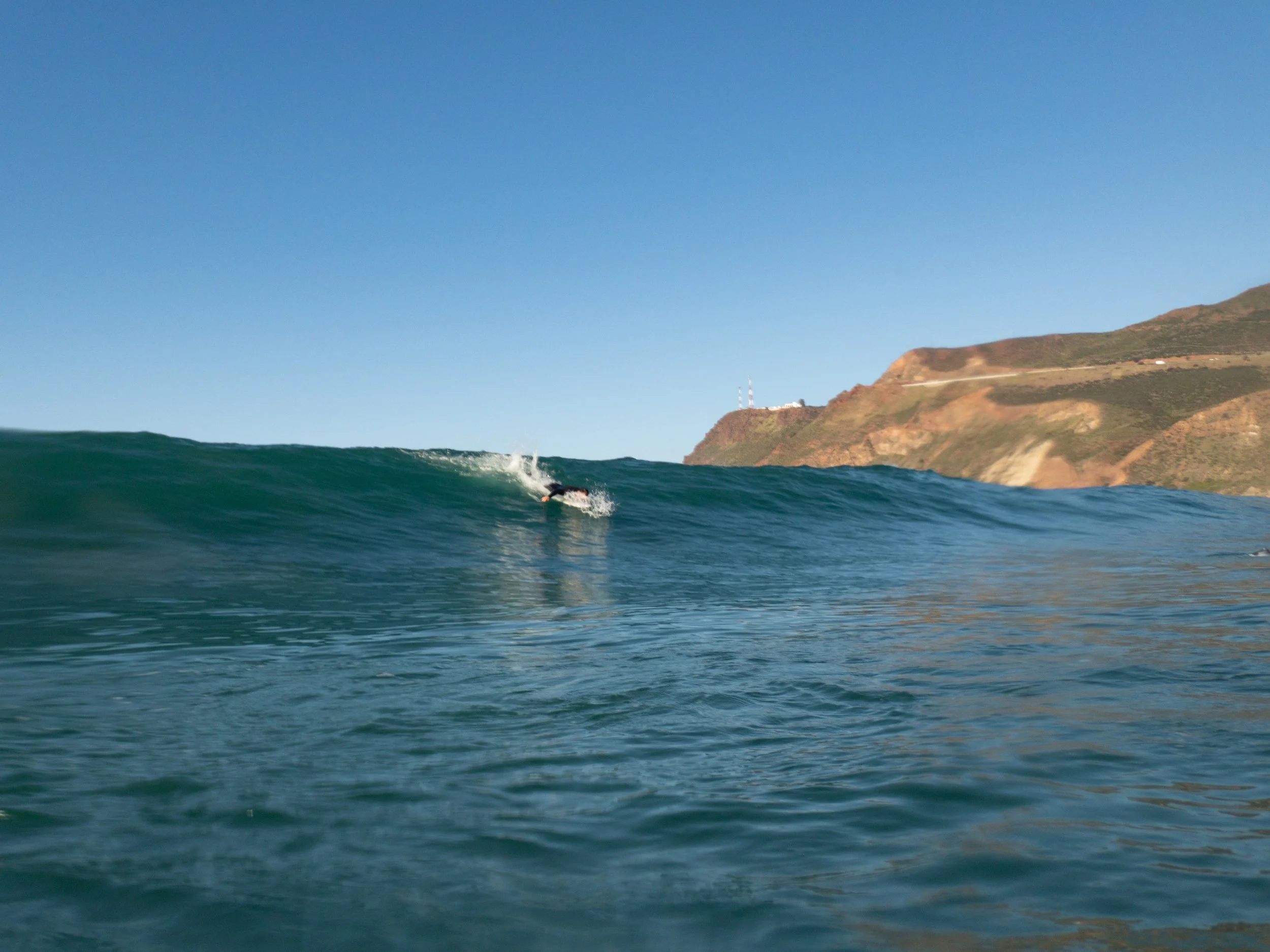 A person surfing on a wave near a coastal mountain landscape under a clear blue sky.
