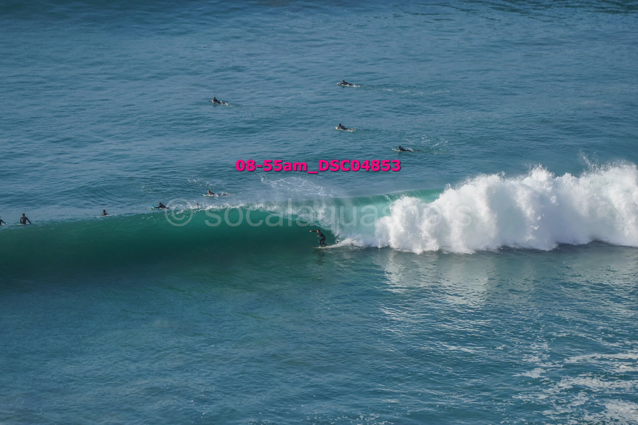 Surfer riding a wave with several paddleboarders in the water nearby.