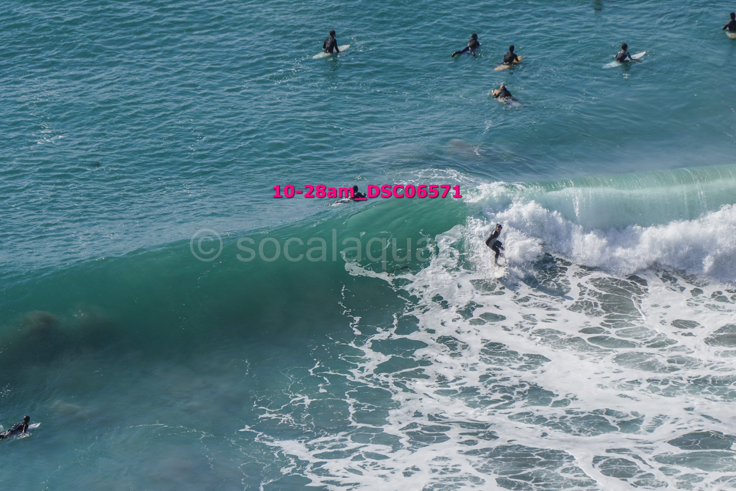 Surfer riding a wave while several surfers wait in the water nearby can be seen in this aerial view of the ocean.