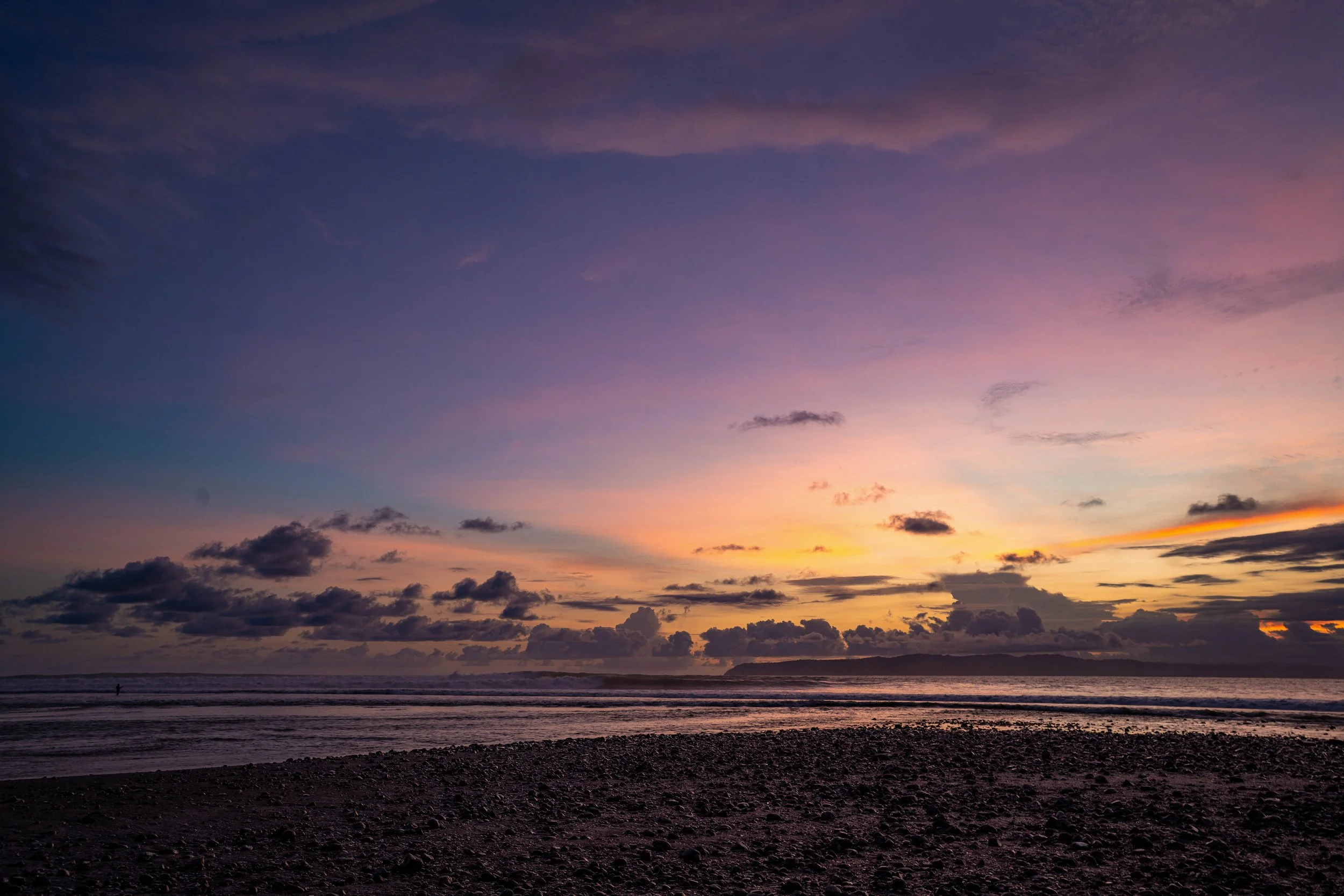 Sunset over the ocean with colorful sky, some clouds, and a person walking on the beach.