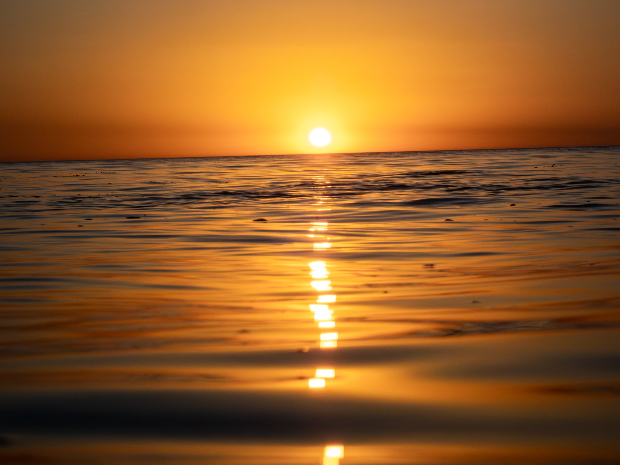 Sunset over calm ocean water with a glowing sun on the horizon and reflection on the water surface.