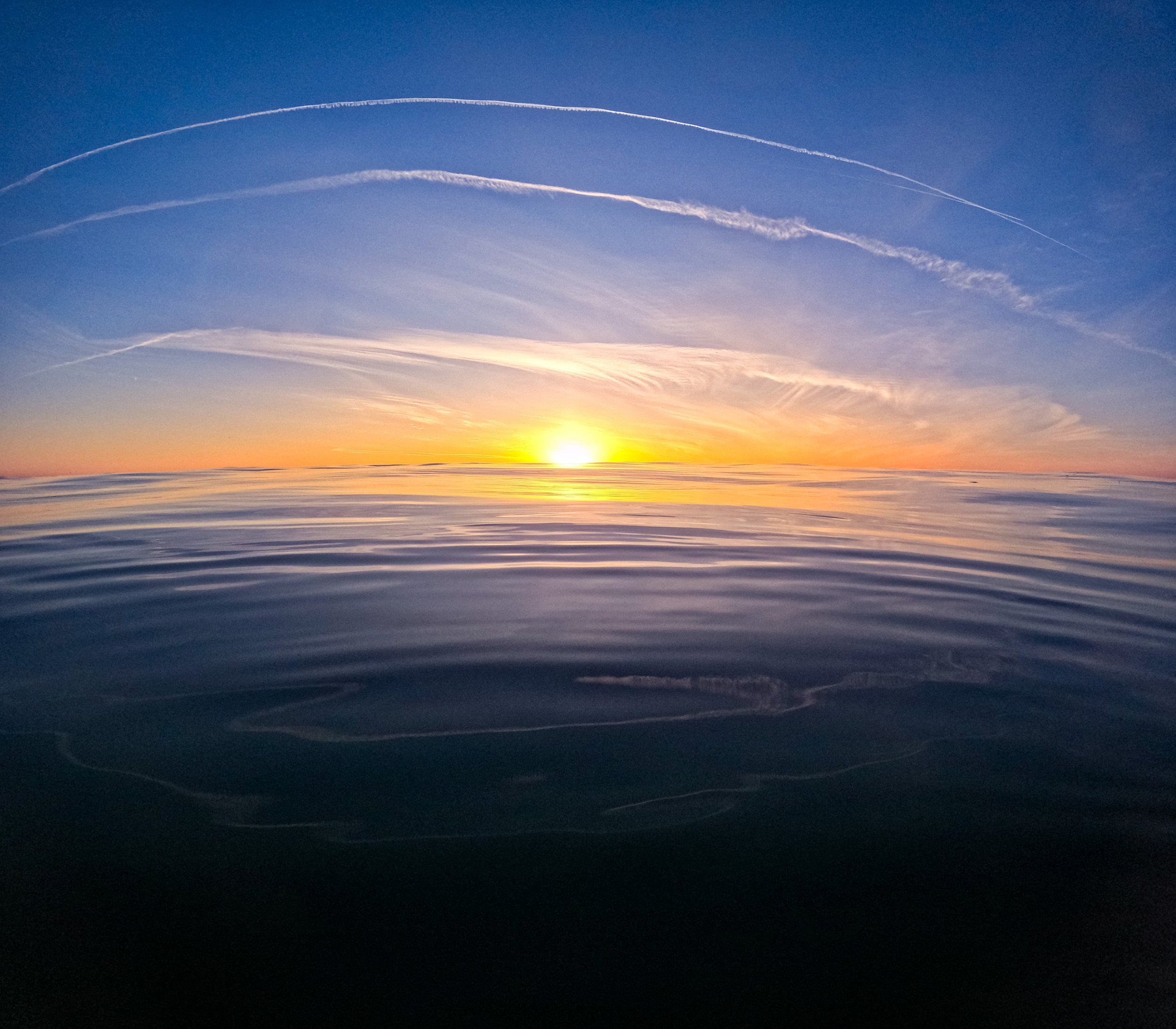 Sunset over calm ocean water with streaks of clouds in the sky.