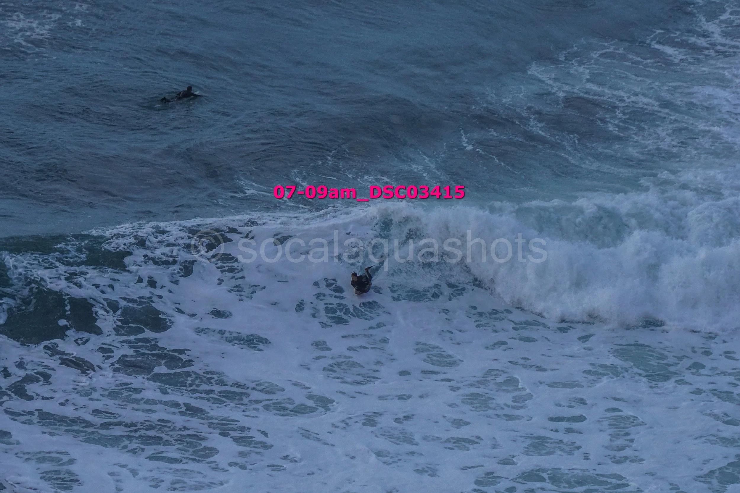 Two surfers in the ocean riding or preparing to ride waves, with one surfer closer to the camera and another further back, amidst white foam and blue water.