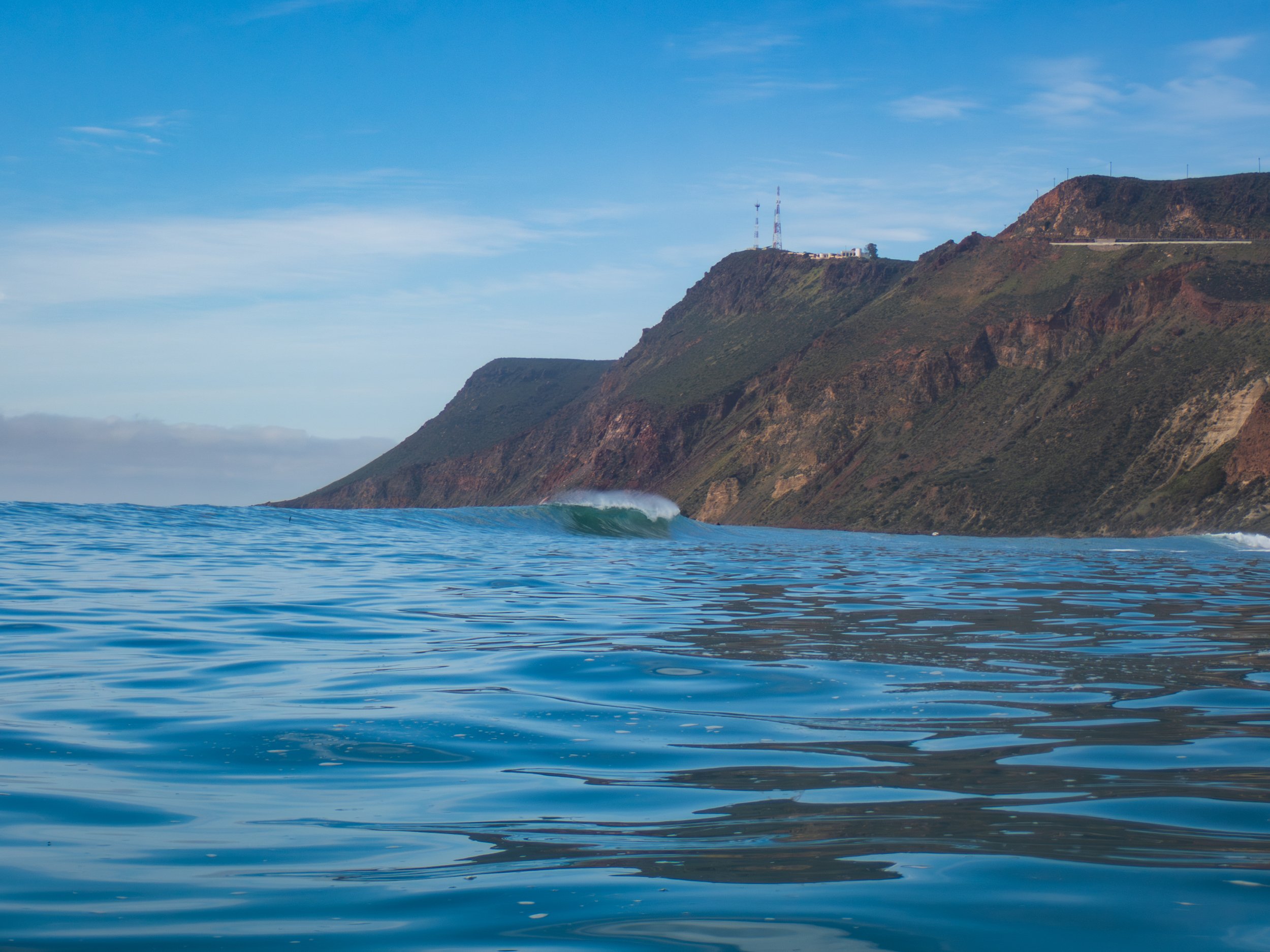Ocean water with small waves in the foreground, hillside with green and brown vegetation, and transmission towers on top of the hill under a partly cloudy blue sky.