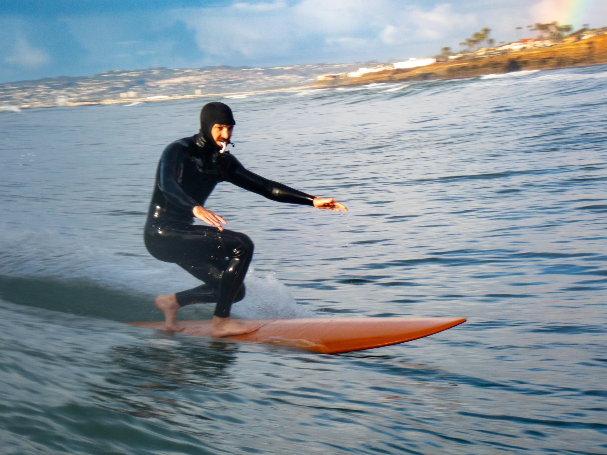 A person wearing a wetsuit and a black hooded cap surfing on a longboard in the ocean during daytime with a coastline and buildings in the background.