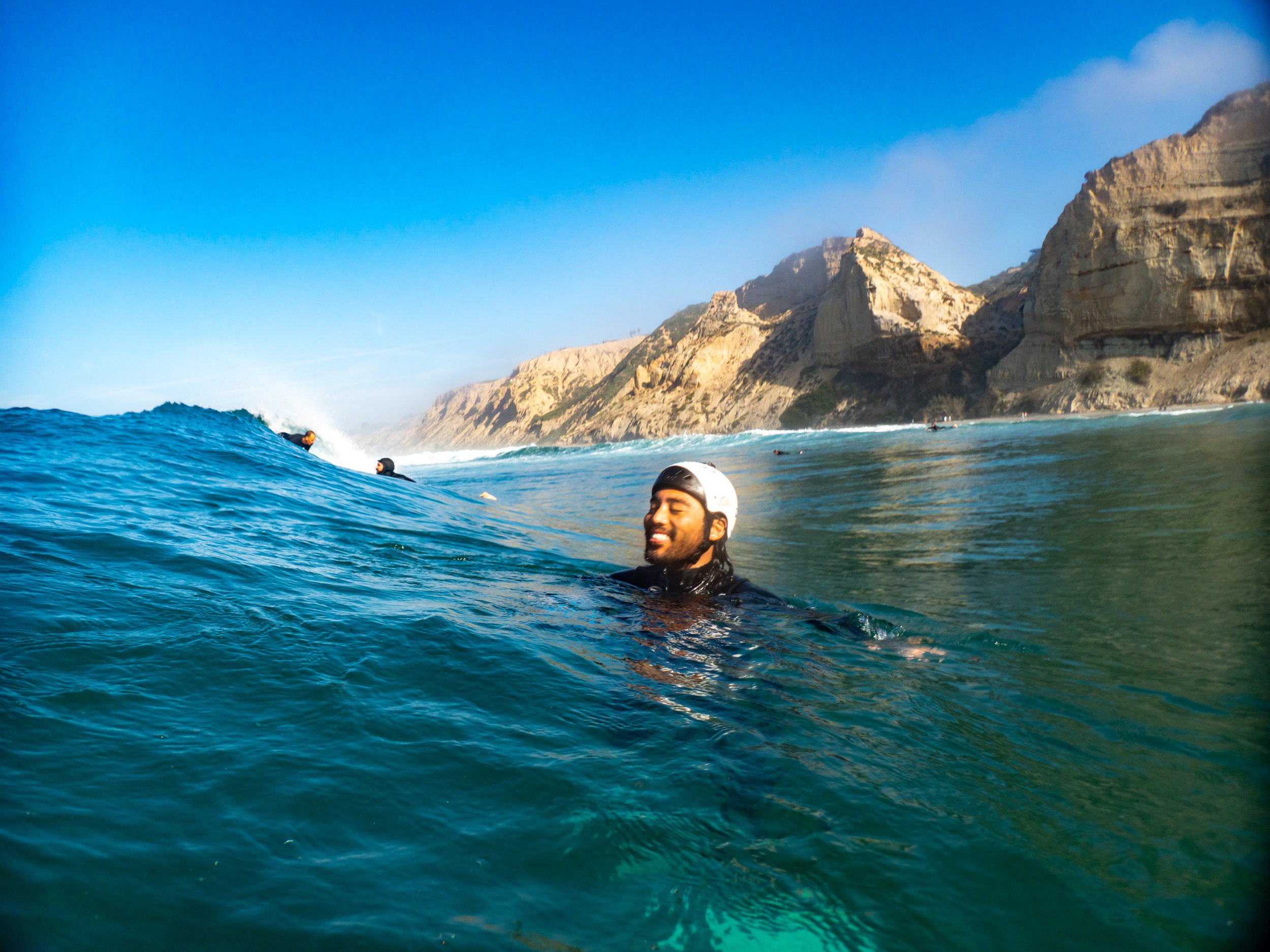 A man in a wetsuit and helmet swimming in the ocean with a smile, surrounded by other surfers and a rocky coastline in the background.