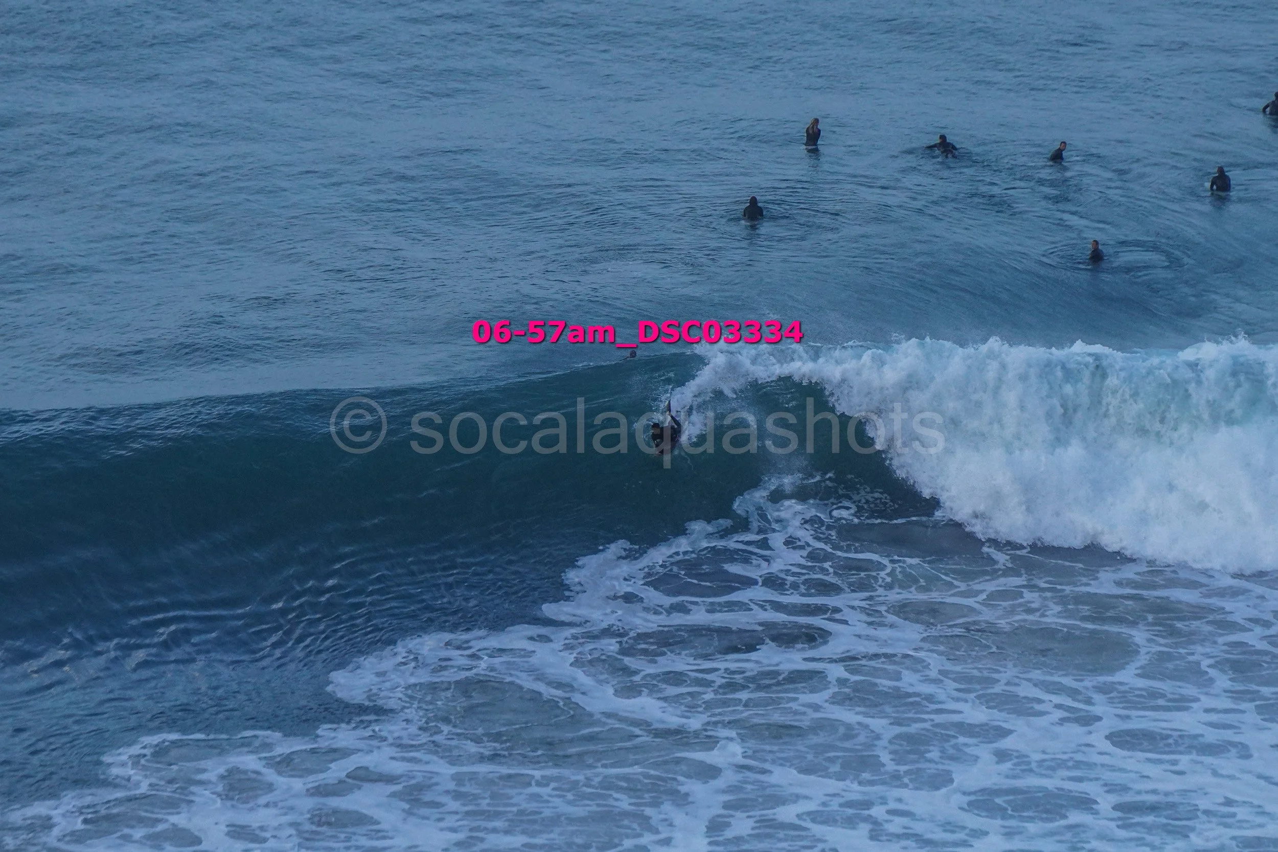 A person surfing on a wave with several people in the water watching, at 6:57 am.