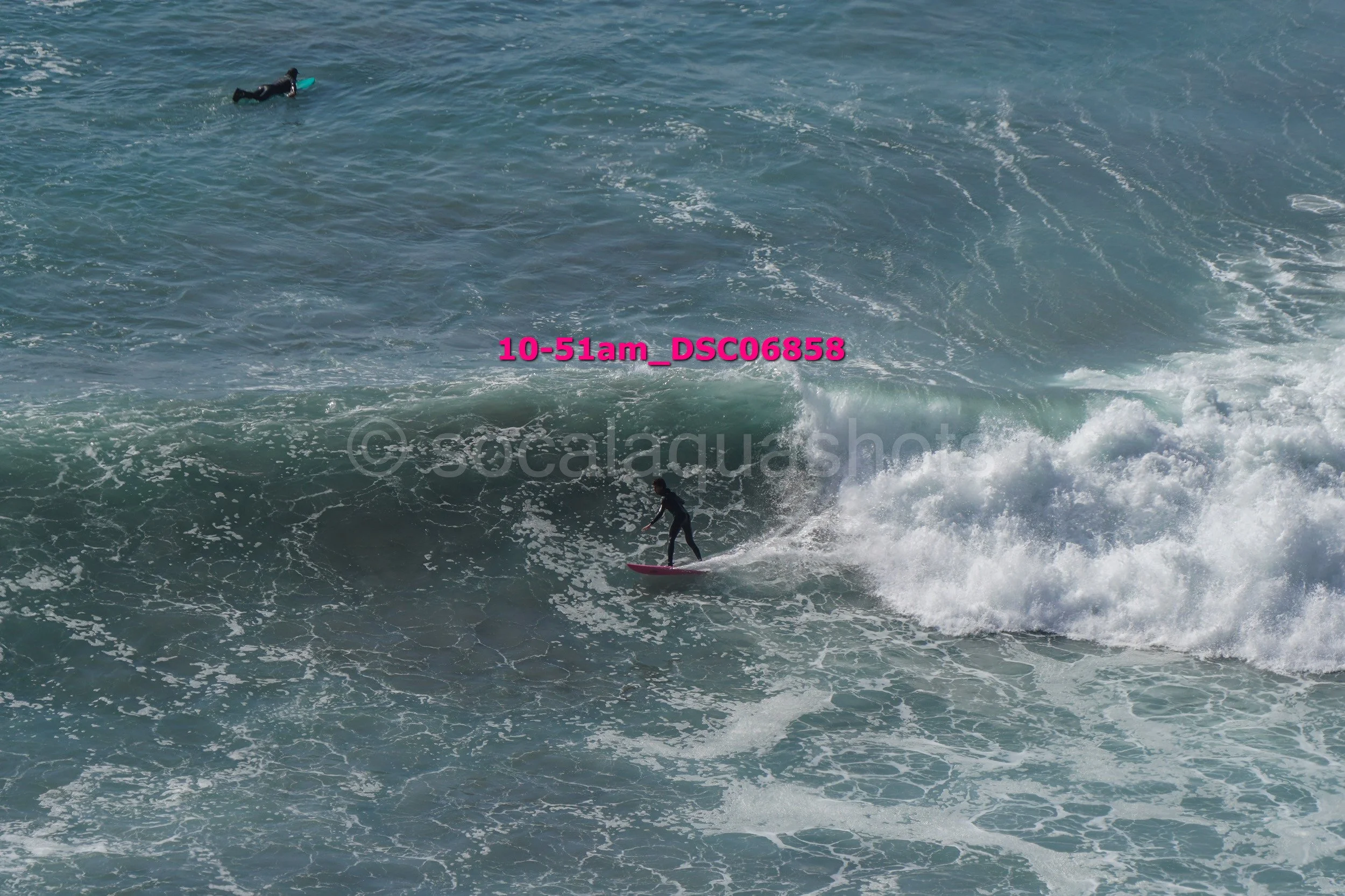 Two surfers riding waves in the ocean, one standing on a pink surfboard and the other lying on a green surfboard.