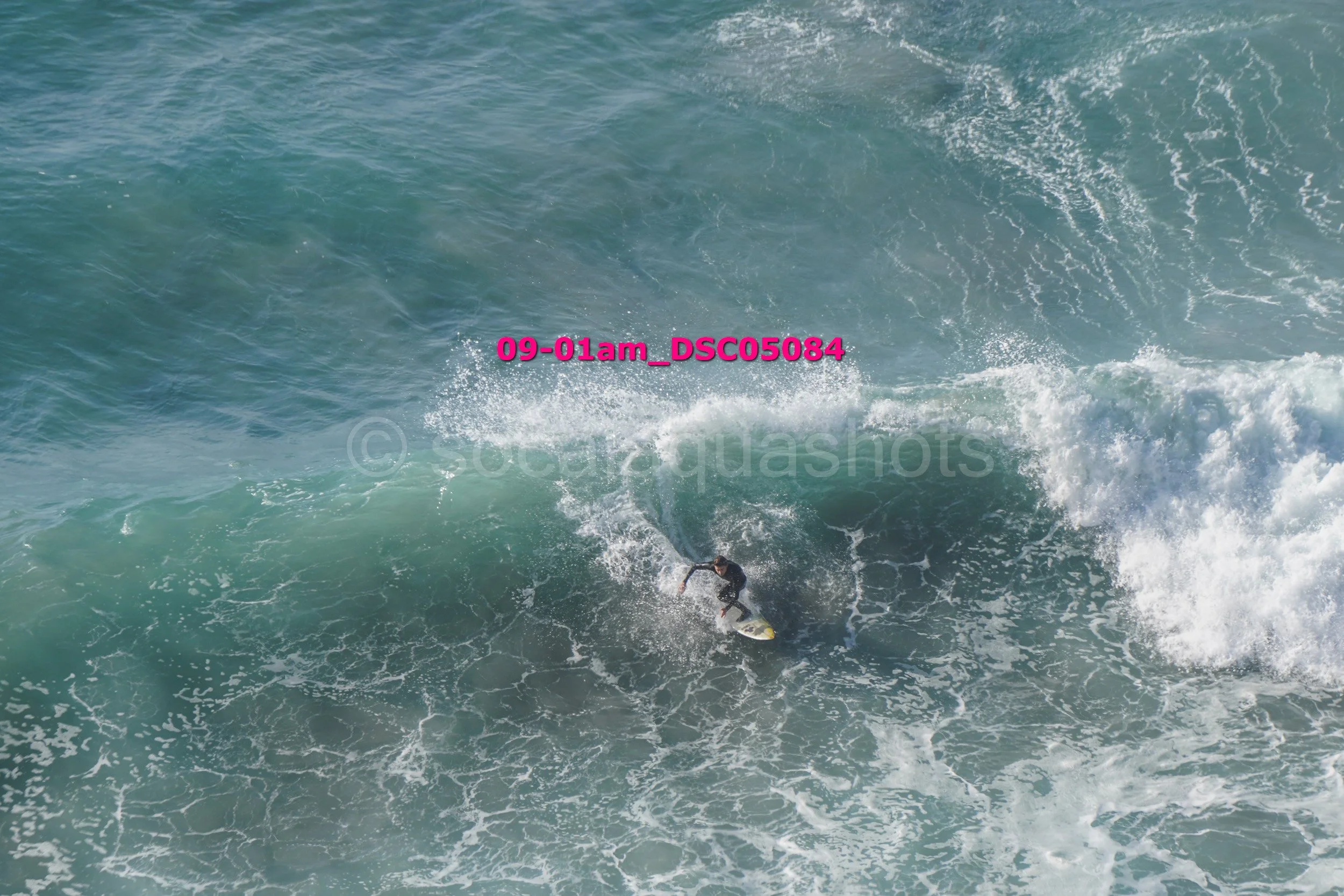 A person riding a surfboard on a large ocean wave, with white foam and water spray around.