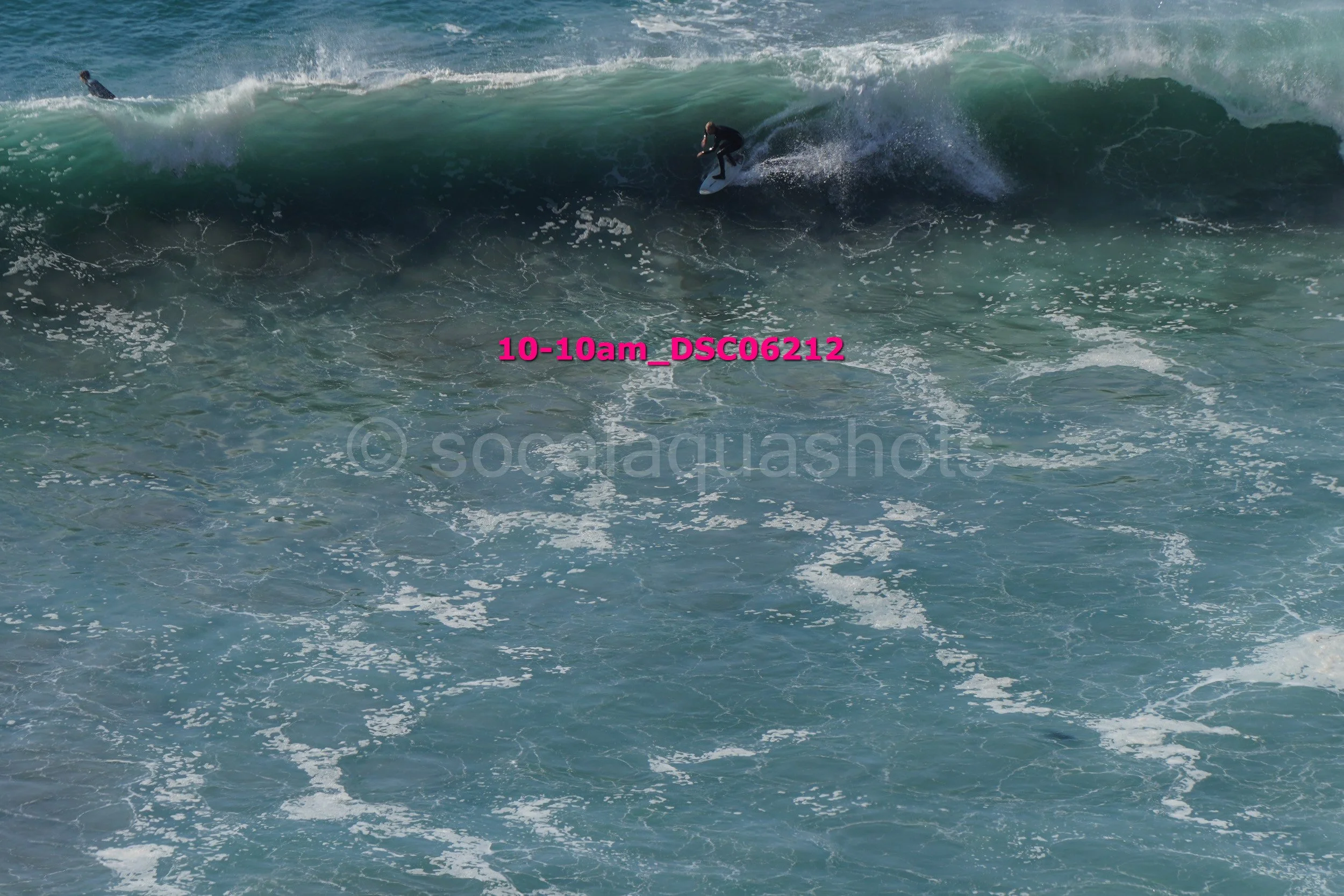Surfer riding a large wave with another surfer visible in the distance, ocean water with foamy white surf, and a watermark 'socialaquashots' across the image.