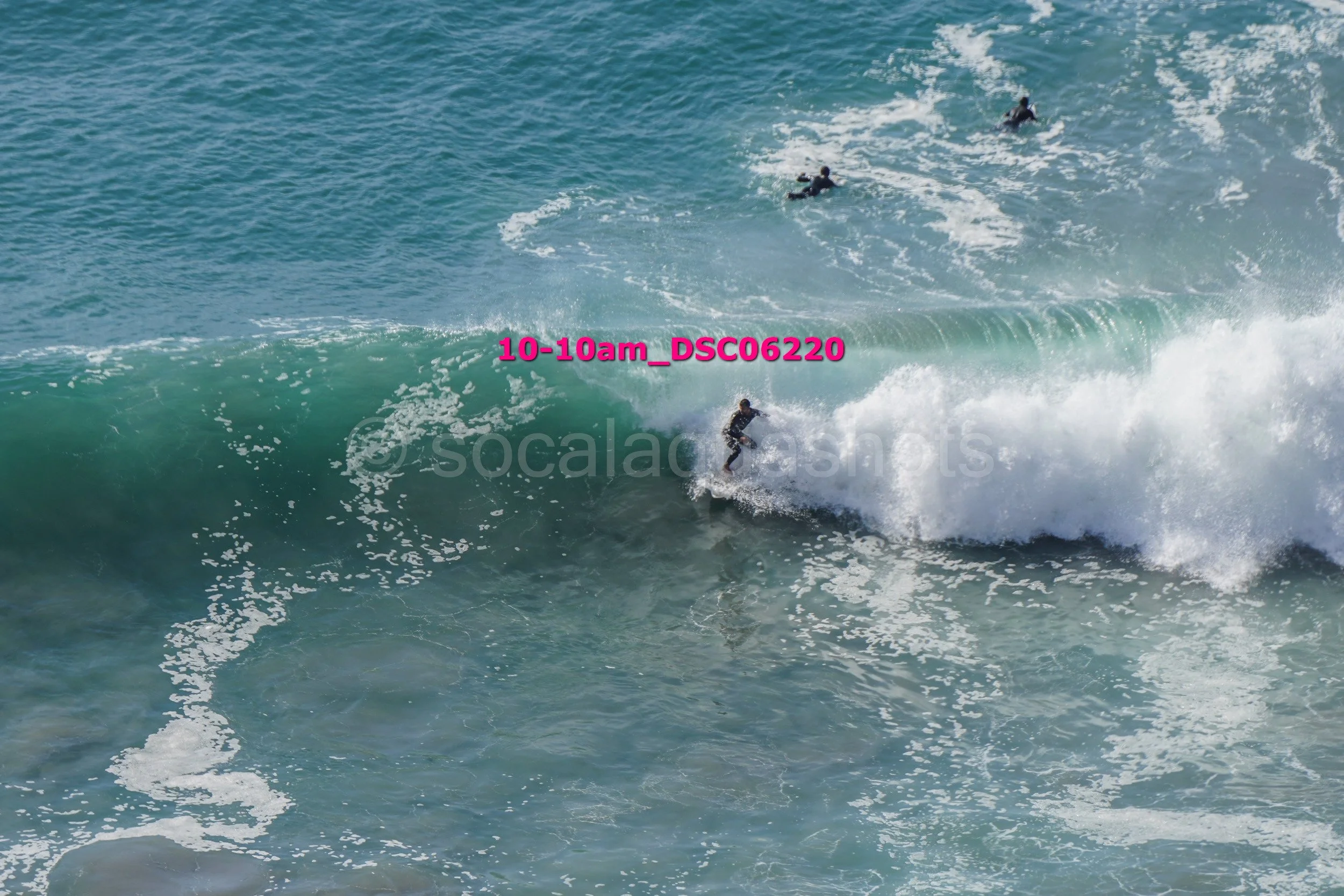 A group of surfers riding a large wave in the ocean, with some surfers farther in the background and one surfer close to the camera riding the wave.