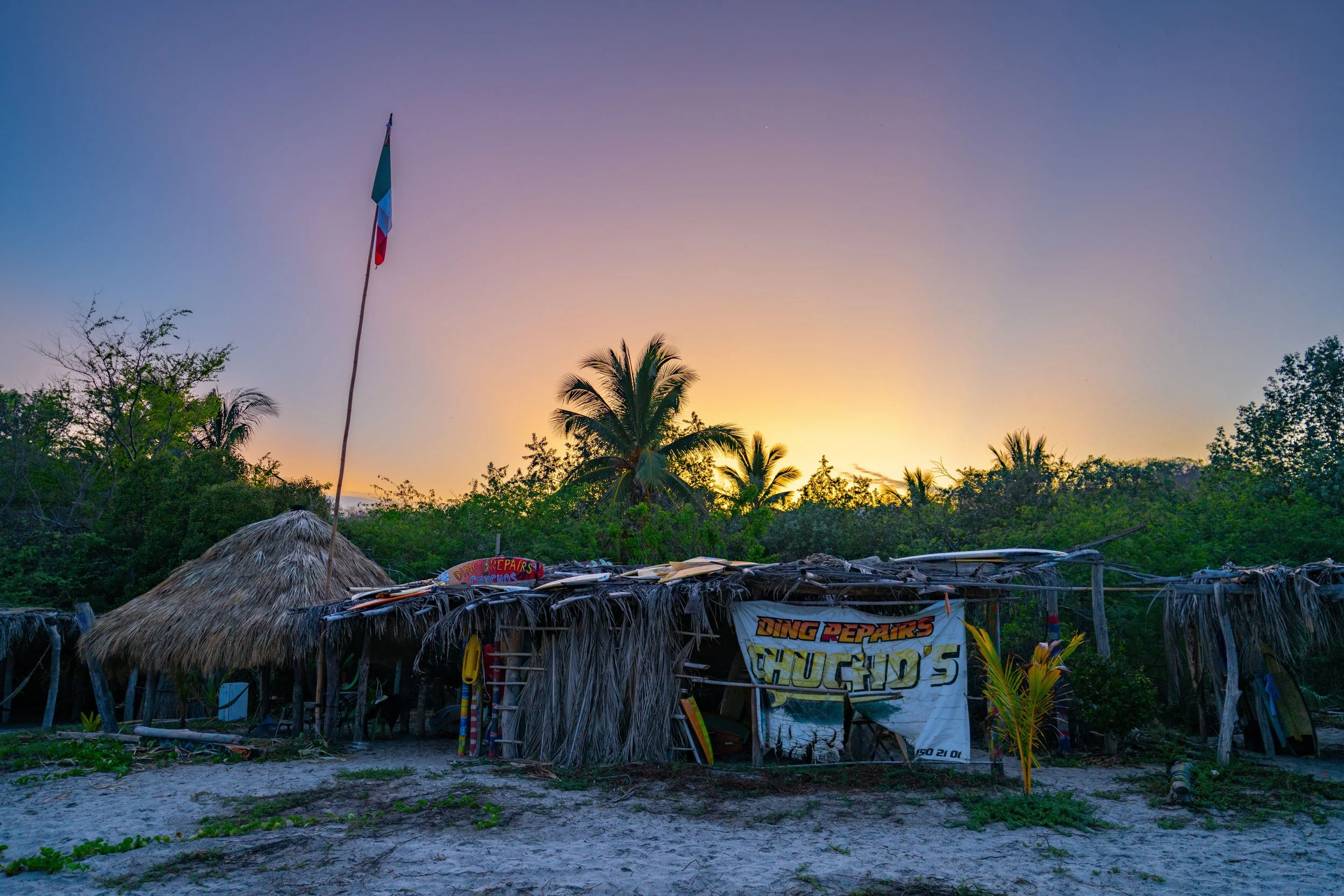 A thatch-roof hut with a sign that reads 'Ding Repairs' and colorful surfboards in front, surrounded by lush green trees with palm trees, and a sunset sky in the background.