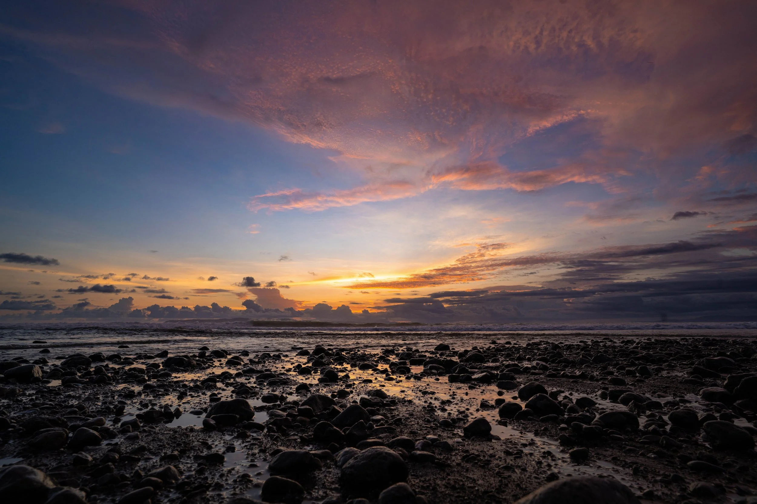 Sunset over the ocean with clouds in the sky and rocks on the beach in the foreground.