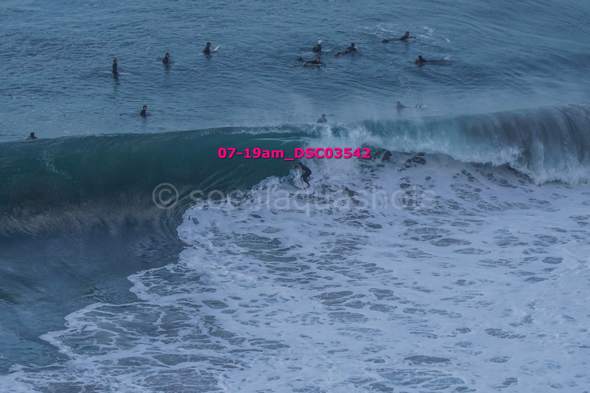 A person surfing on a large wave in the ocean with multiple surfers watching from the water.