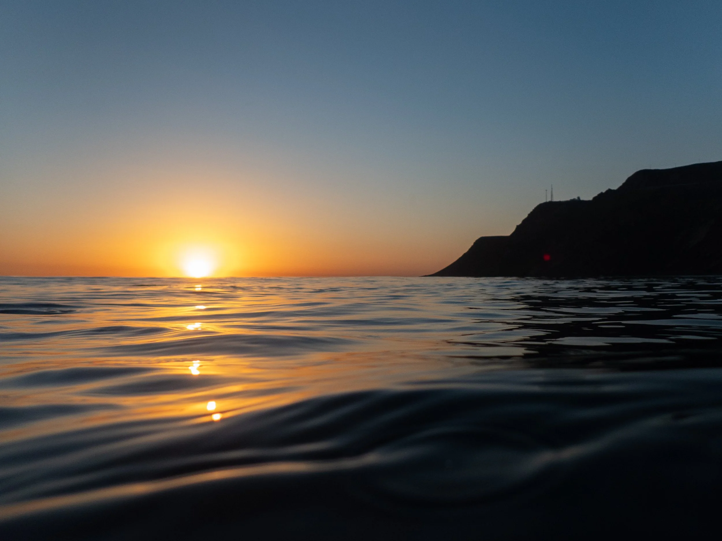 Sunset over the ocean with calm water reflecting the orange and yellow sky, and a dark coastline on the right.