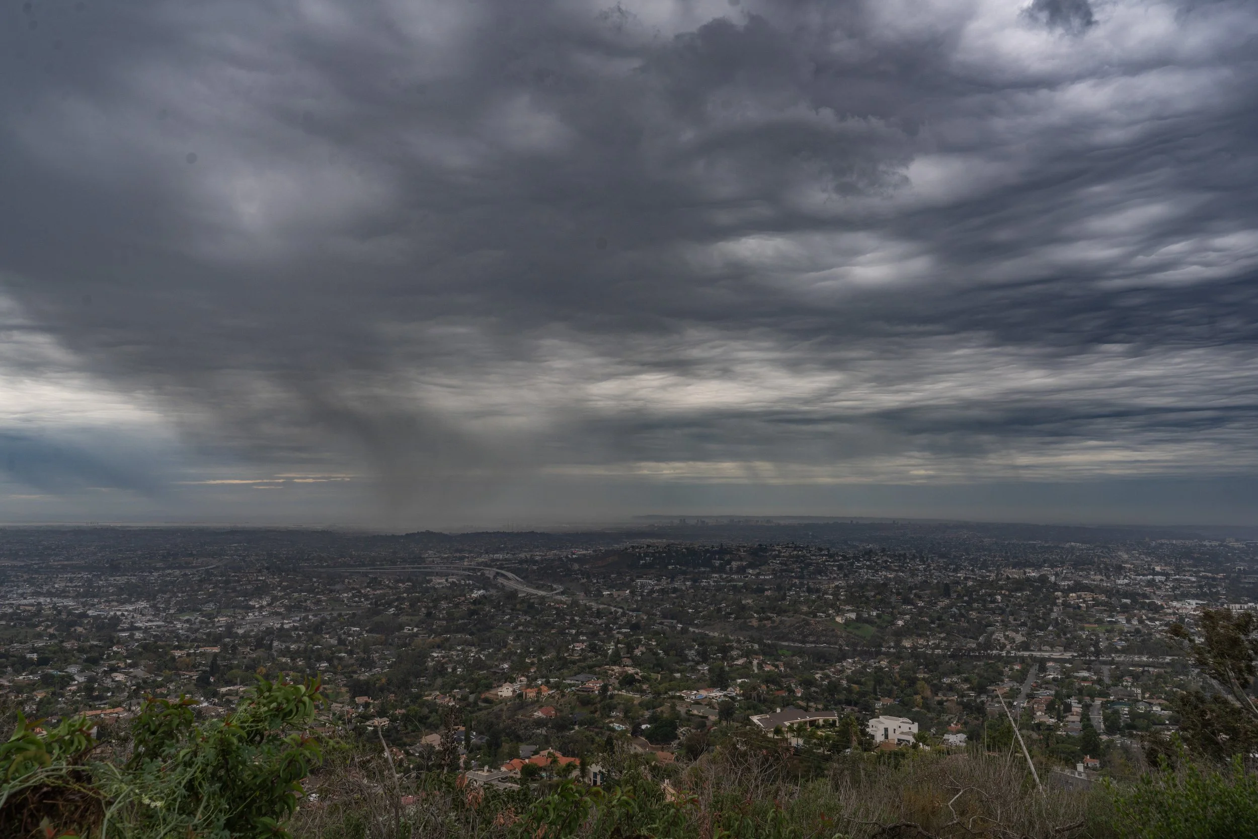 A cityscape under a dark, cloudy sky with rain falling in the distance, taken from a hillside or mountain.
