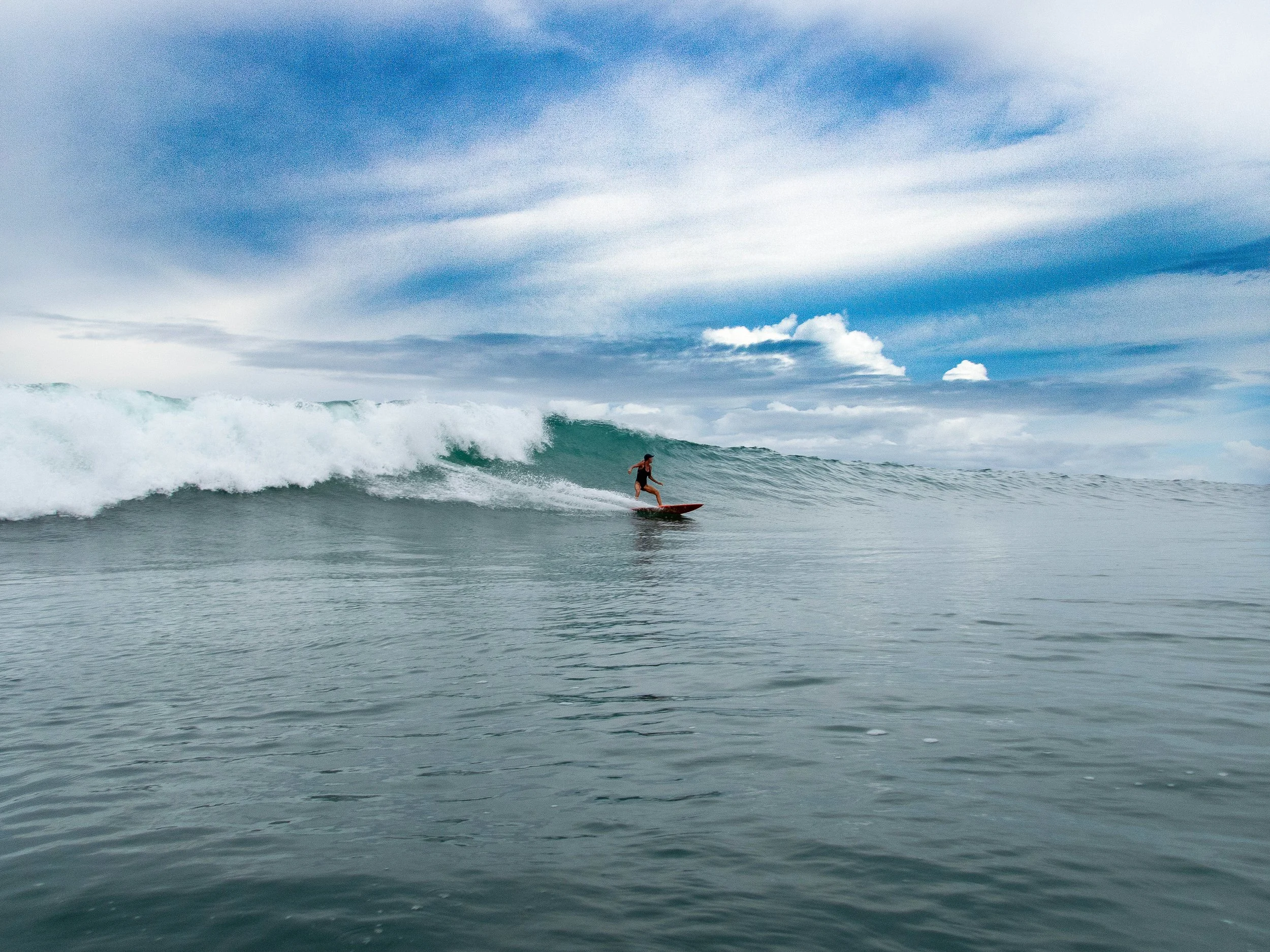 Surfer riding a large wave under a cloudy blue sky.