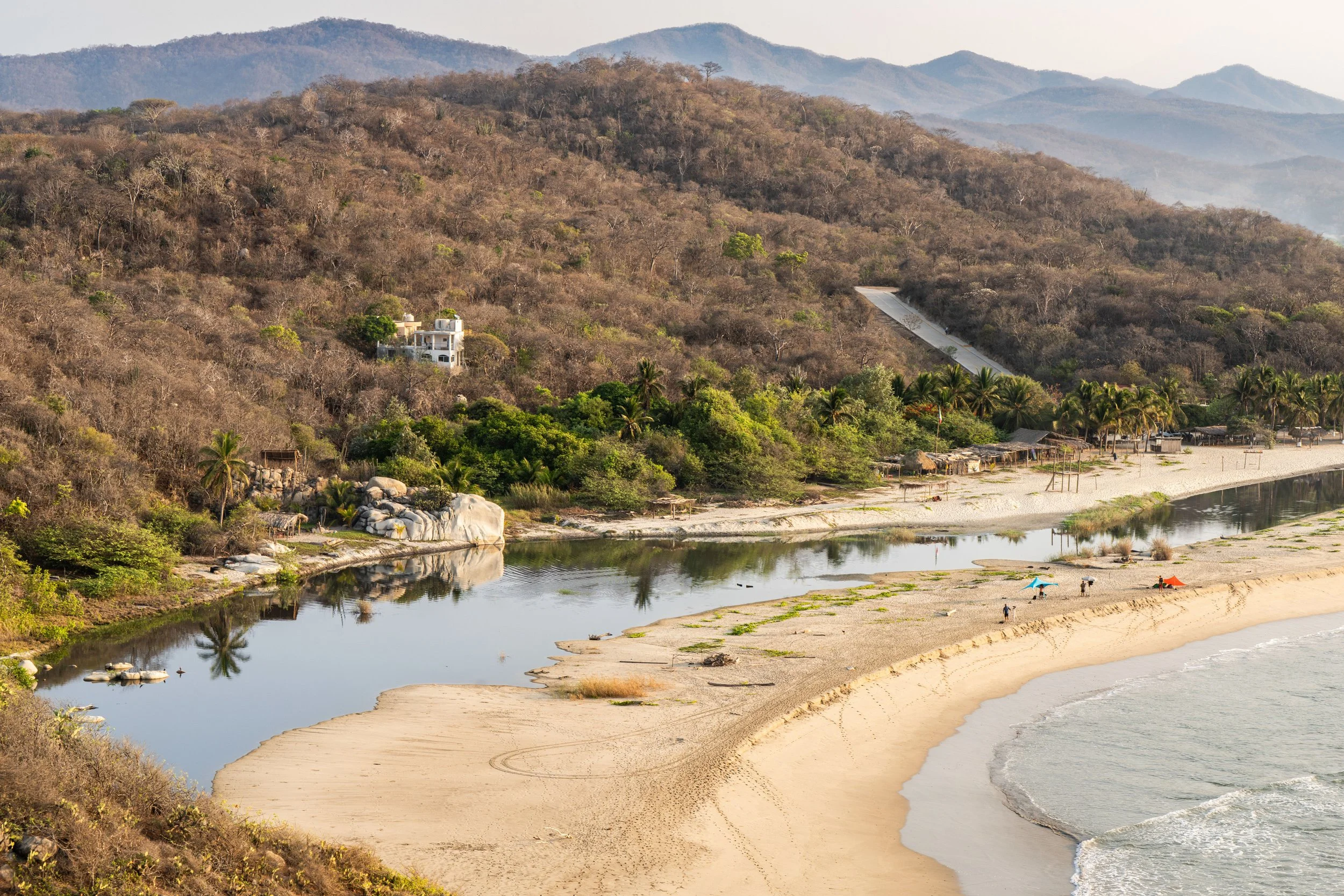 A scenic view of a beach with calm water, sandy shores, and a few umbrellas. In the background, there are green trees, a hillside, and mountain ranges under a slightly cloudy sky.