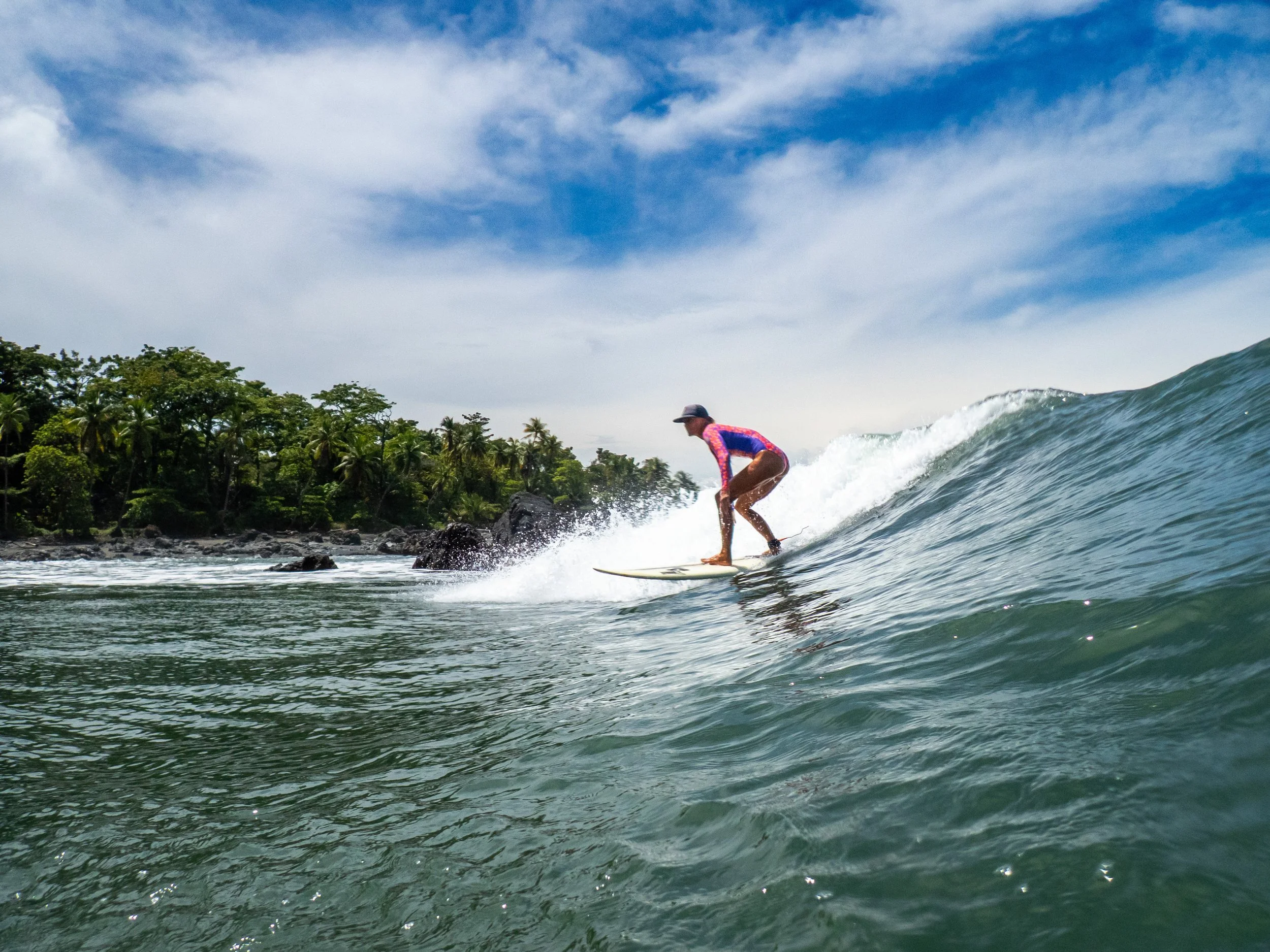 A person surfing on a wave near the shoreline with lush green trees and a partly cloudy sky in the background.