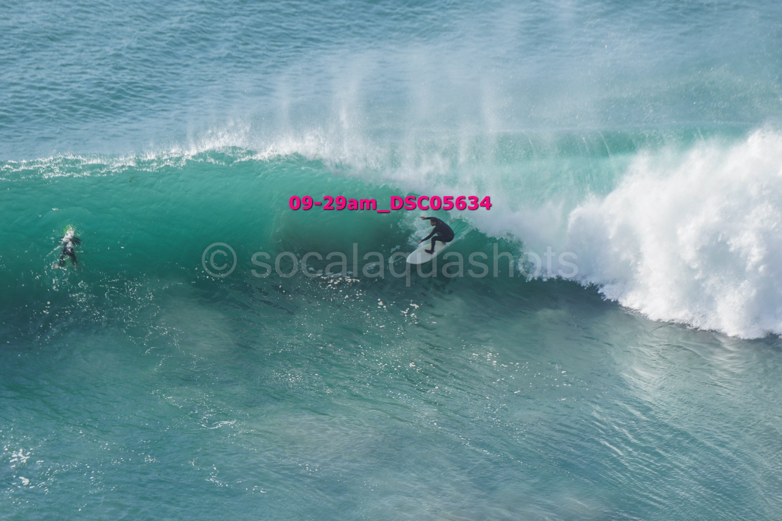 A person riding a surfboard on a large ocean wave with another surfer in the distance.