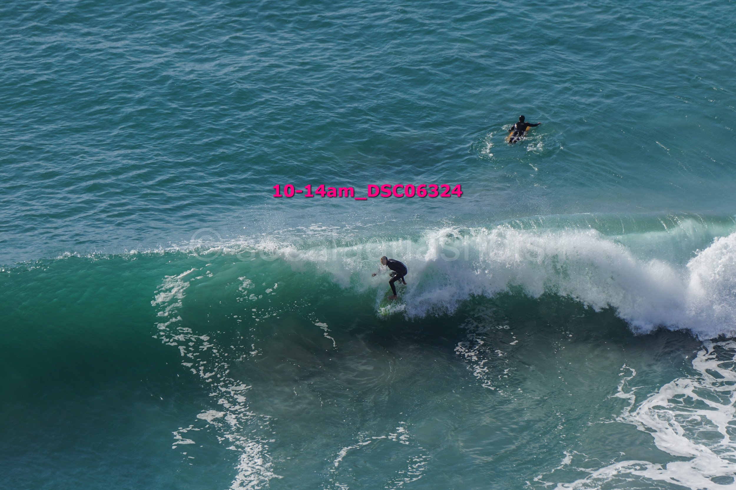 A person surfing on a green wave in the ocean, with another person swimming behind them.