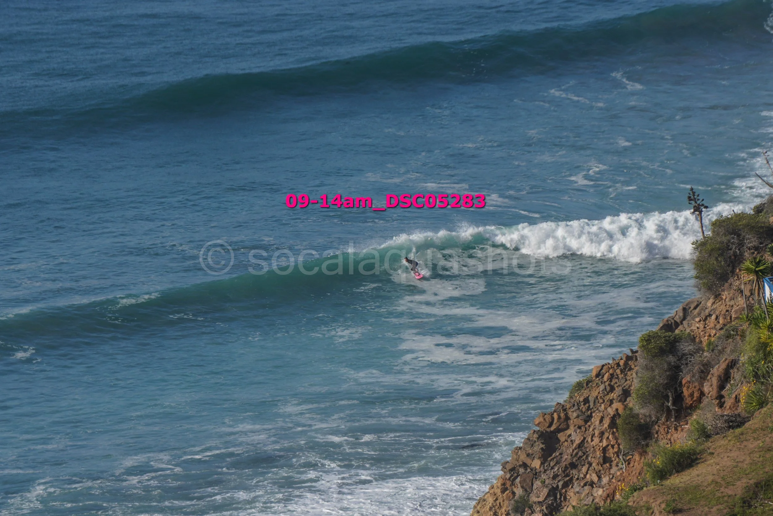 A person surfing on a wave near a rocky coastline with some vegetation.